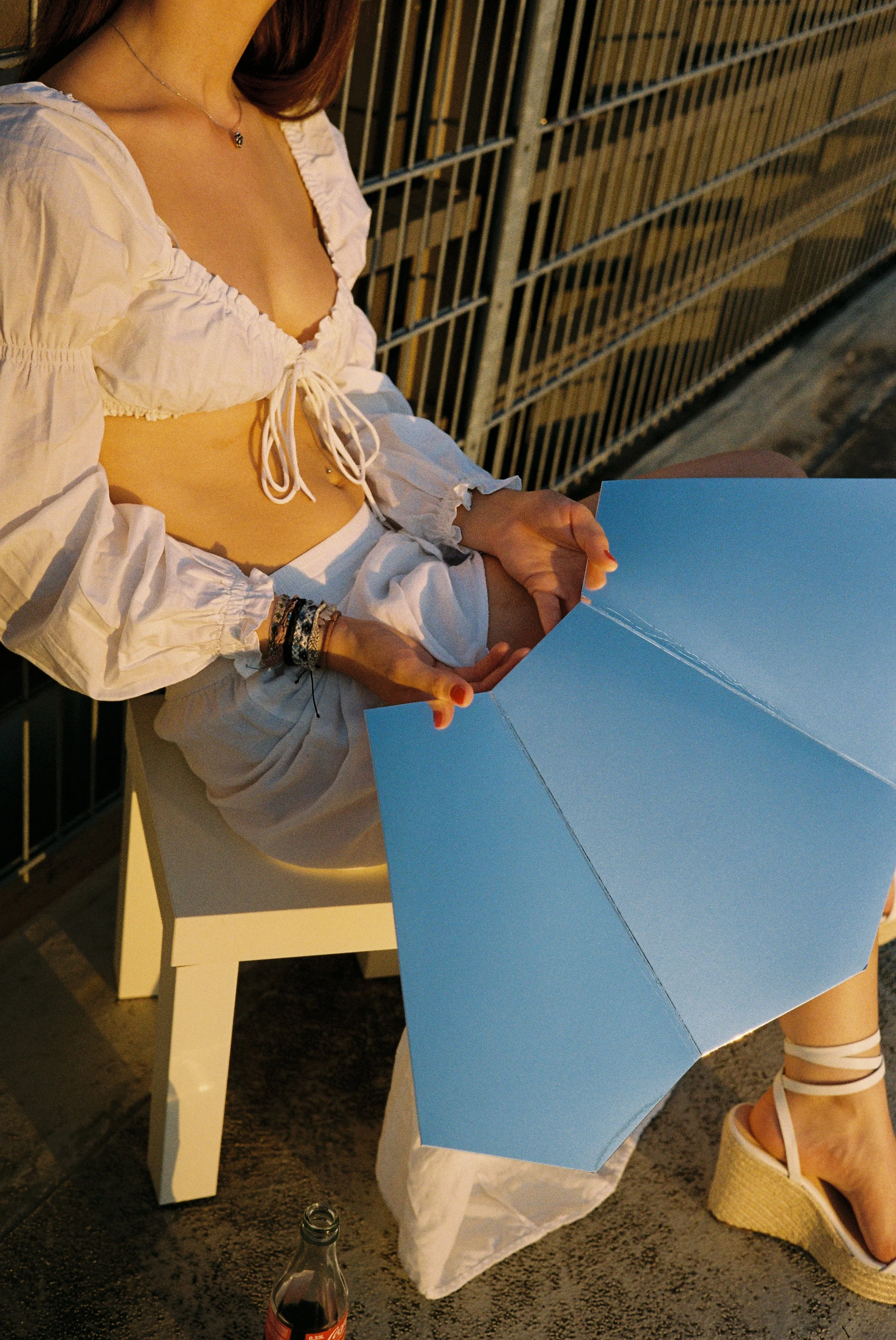 Eine junge Frau sitzt auf einem Stuhl auf einer Terrasse bei Sonnenuntergang und hält einen blauen Regenschirm in der Hand. Sie trägt ein weißes, lockeres Top und weiße Hosen sowie weiße Keil-Sandaletten.