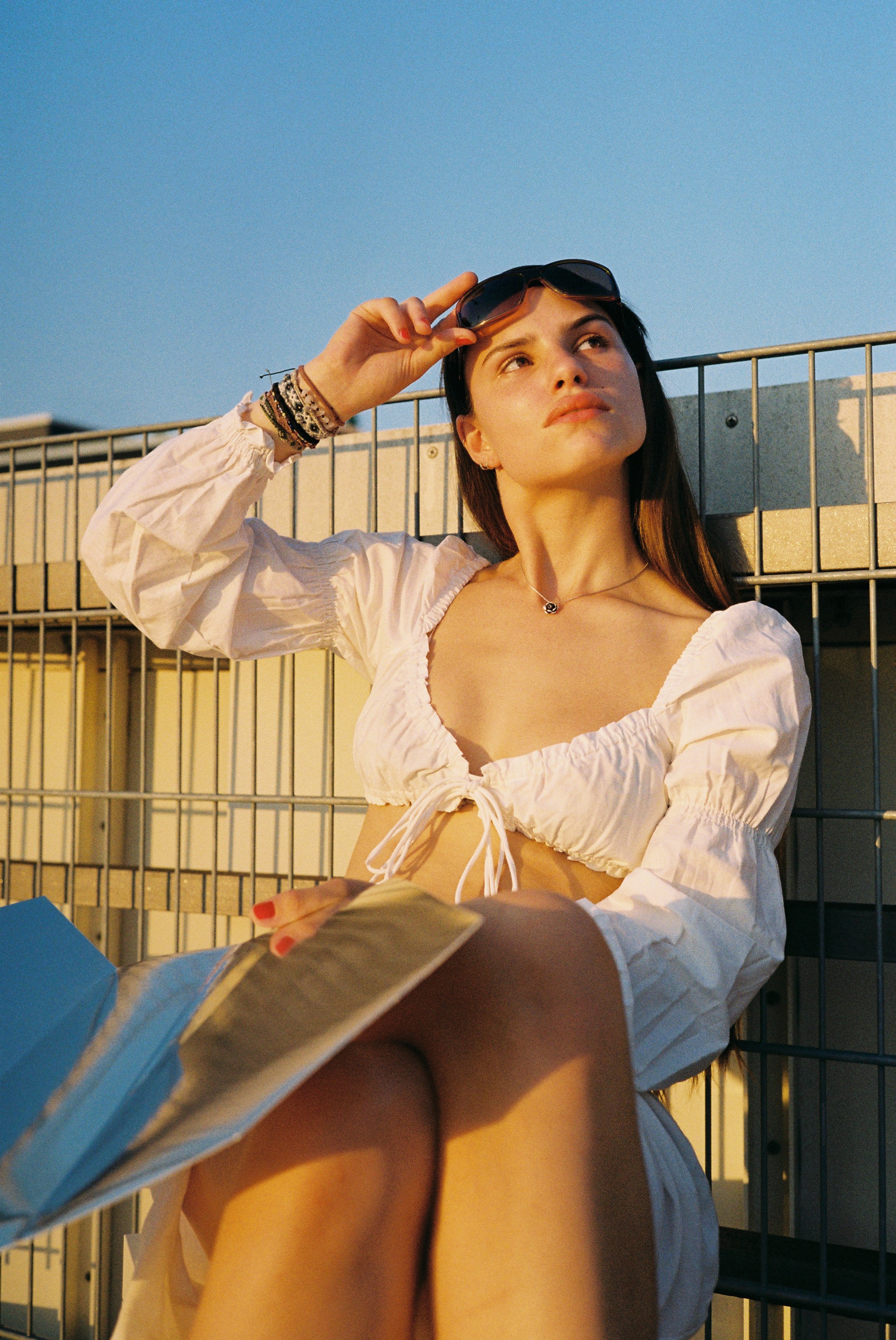 Junge Frau mit dunklen Haaren sitzt vor einer Metallzaun im Sonnenlicht, trägt eine weiße Bluse und Sonnenbrille, liest ein Magazin.