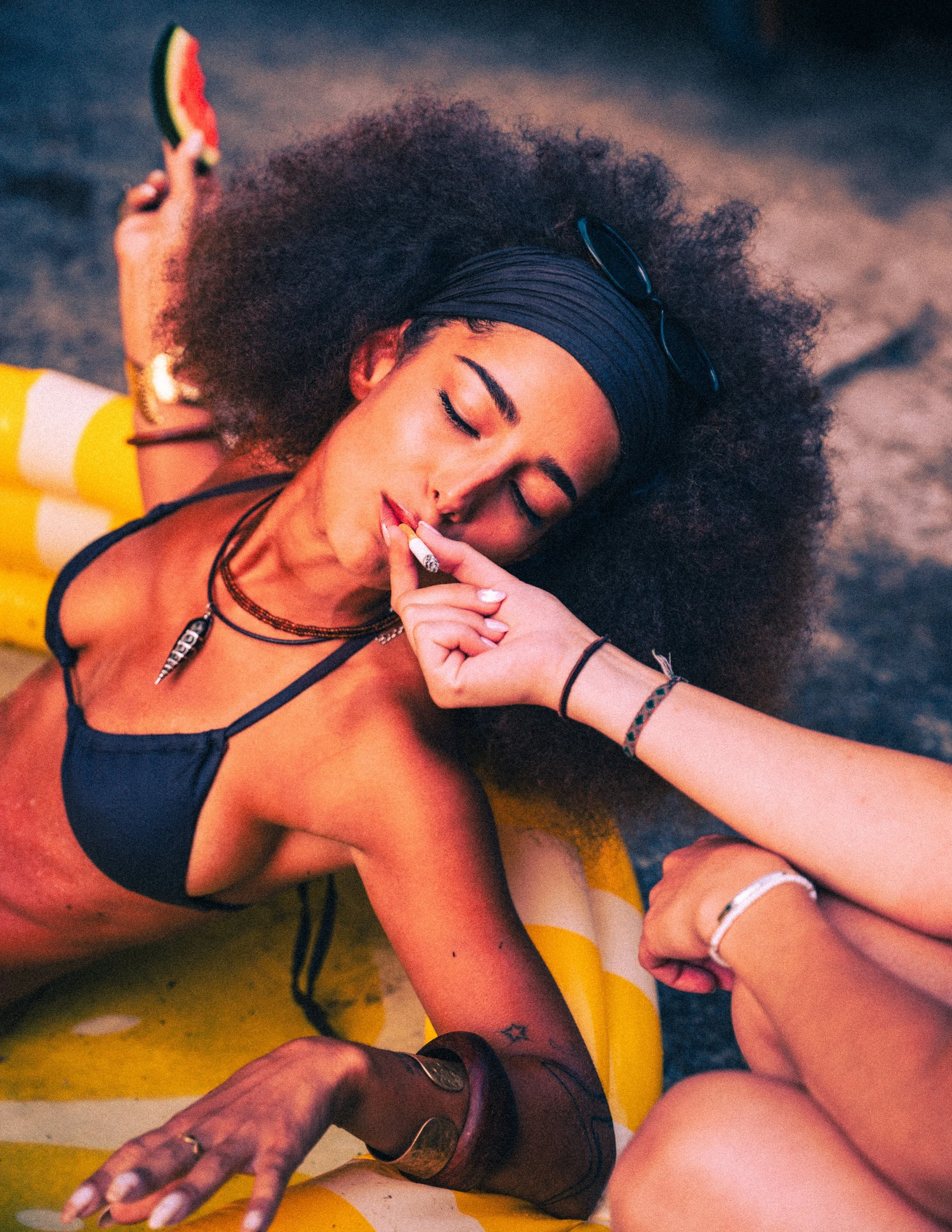 Eine junge Frau mit lockigem Haar, Sonnenbrille auf dem Kopf, liegt auf einer Luftmatratze im Sand, trägt einen schwarzen Bikini und raucht eine Zigarette.