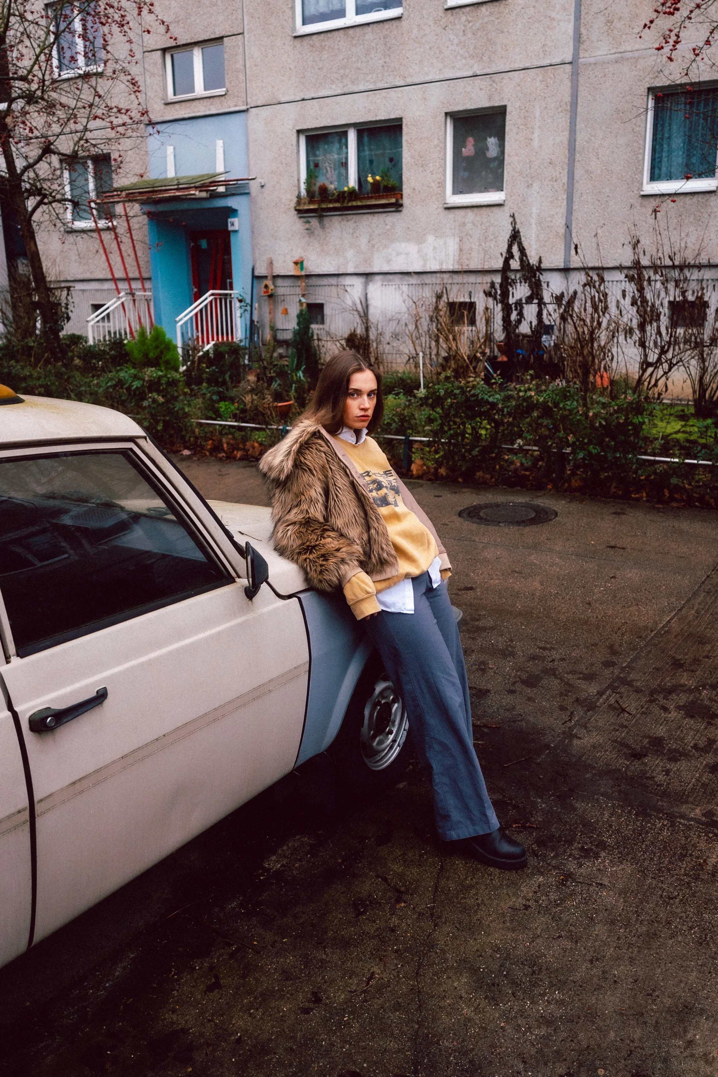 Eine junge Frau lehnt an einem alten Auto auf einem Asphalt. Im Hintergrund sind ein Mehrfamilienhaus und ein Garten mit verwelkten Pflanzen zu sehen.