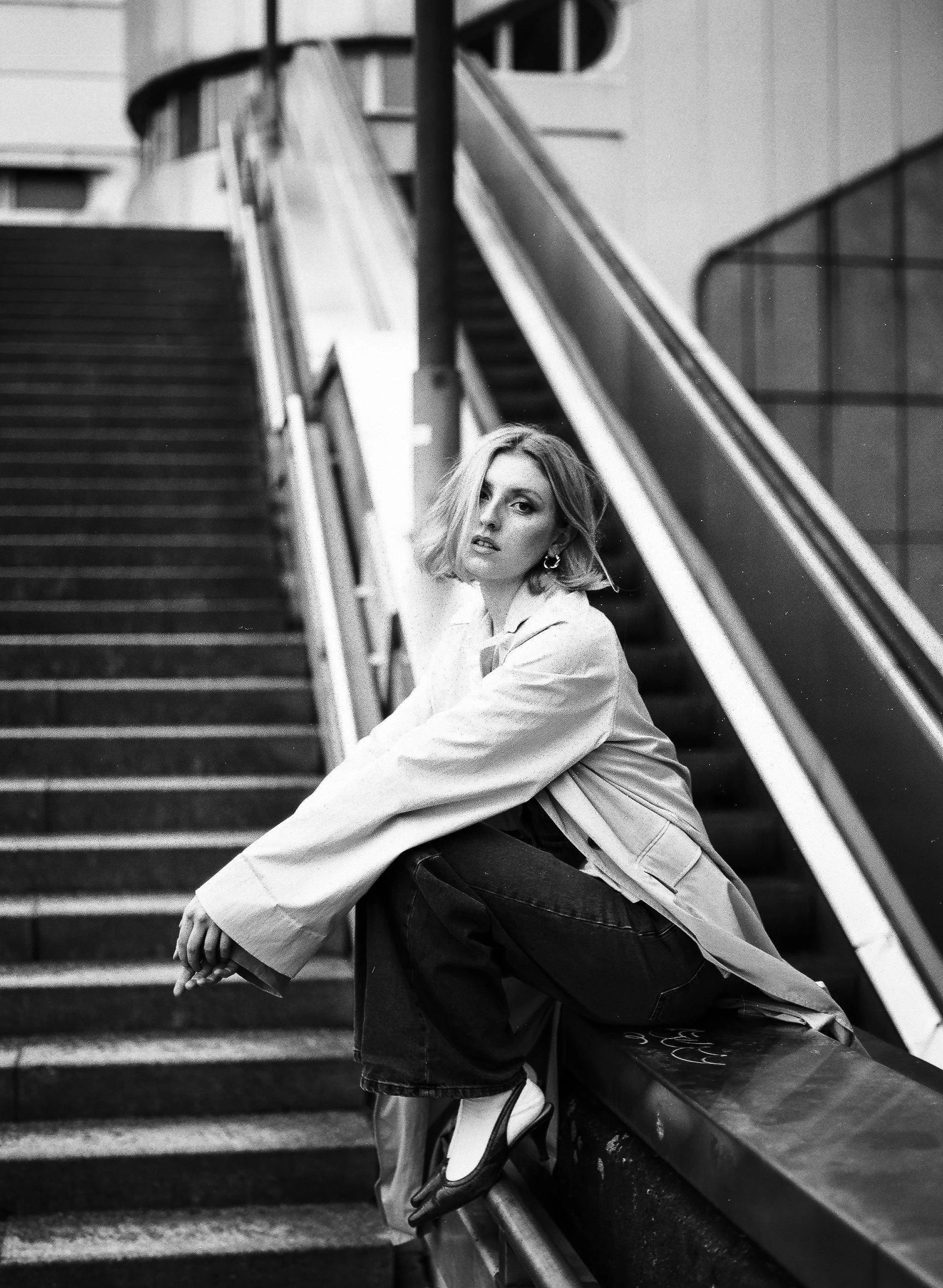 Schwarz-Weiß-Fotografie einer jungen Frau mit kurzen, lockigen Haaren, die auf einer Treppe in einer urbanen Umgebung sitzt. Sie trägt eine helle Jacke und dunkle Hosen, schaut nachdenklich in die Kamera.