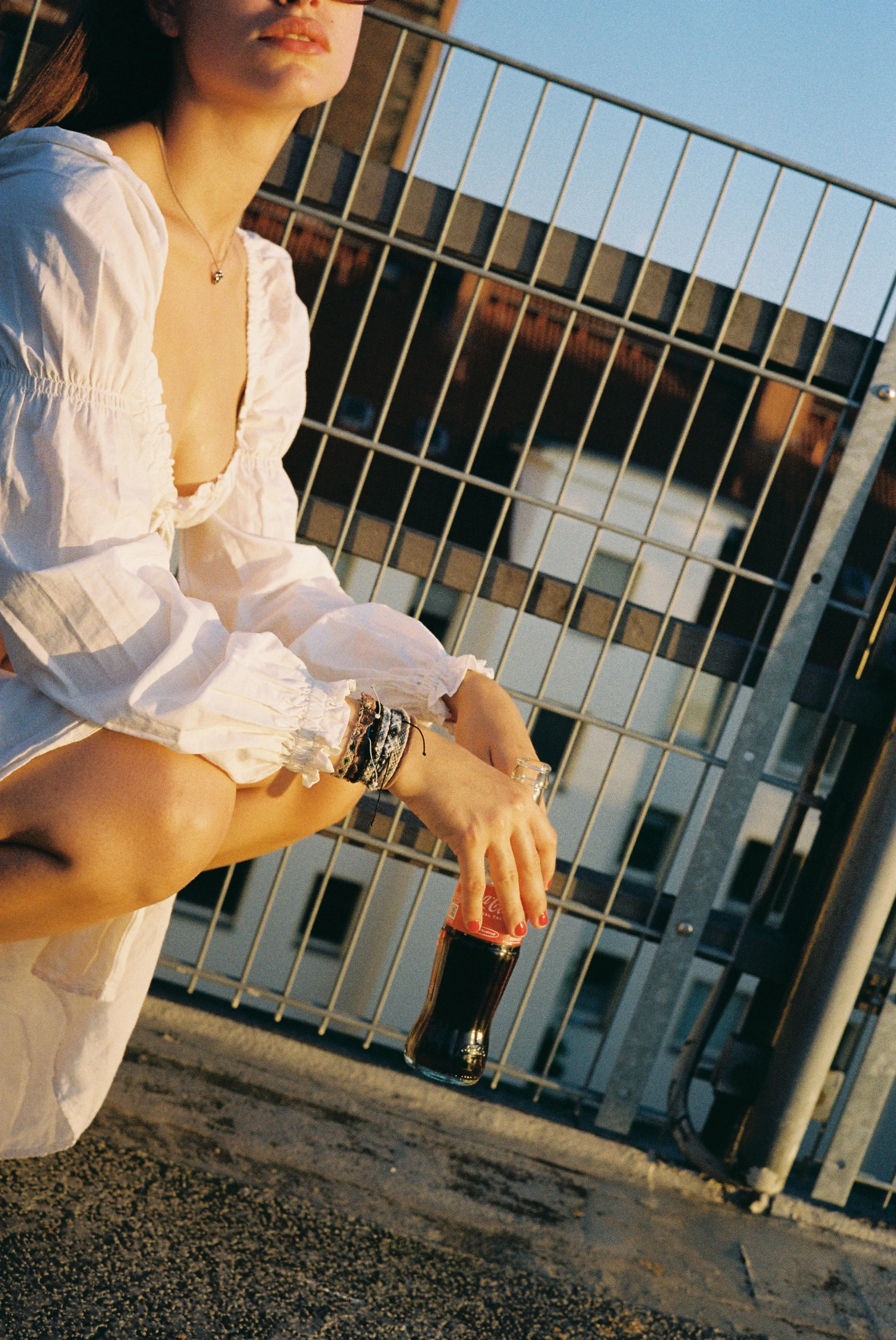 Eine junge Frau sitzt auf dem Boden einer Terrasse oder eines Daches, die Sonne scheint hell. Sie trägt ein weißes, gewickeltes Oberteil mit Puffärmeln und mehrere Armbänder. Sie hält eine Flasche Cola in der Hand.