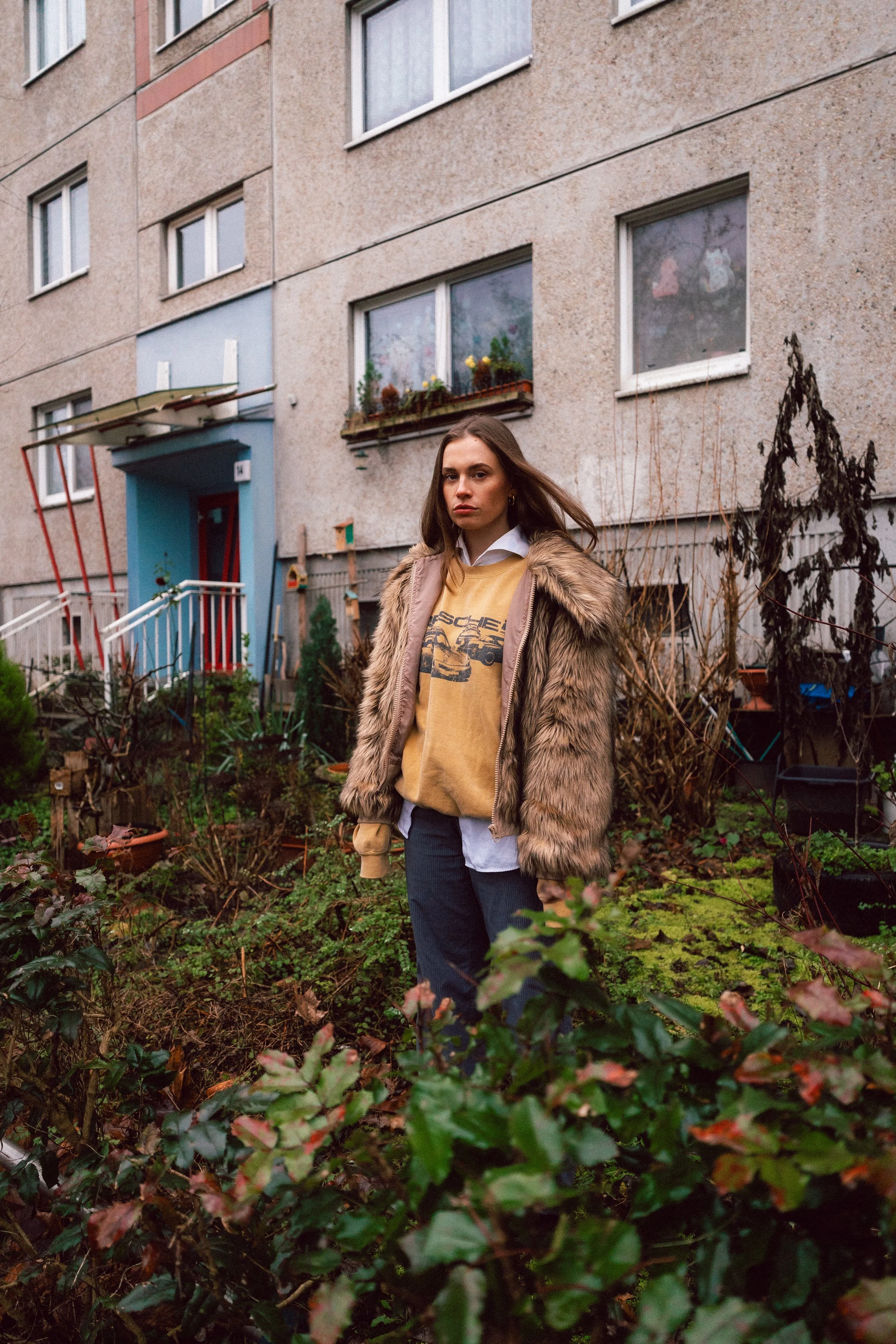 Eine junge Frau steht in einem Garten vor einem Mehrfamilienhaus mit einem braunen Fassaden und mehreren Fenstern, umgeben von Pflanzen und Büschen.