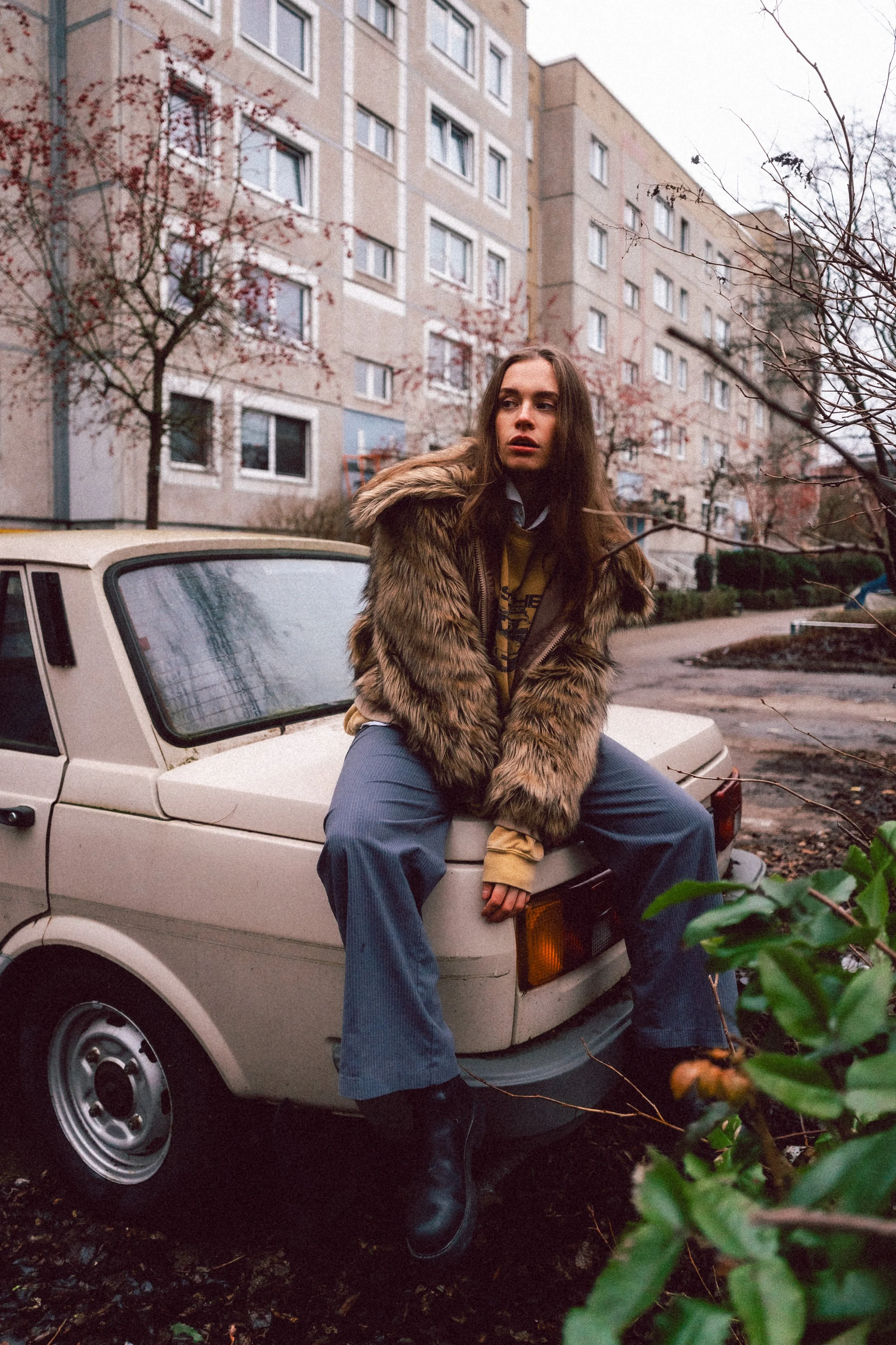 Eine junge Frau sitzt auf dem vorderen Teil eines weißen Autos vor einem mehrstöckigen Apartmentgebäude, umgeben von Büschen und Bäumen, bei bewölktem Himmel, mit langem, braunem Haar, einem pelzähnlichen Mantel und Jeans.
