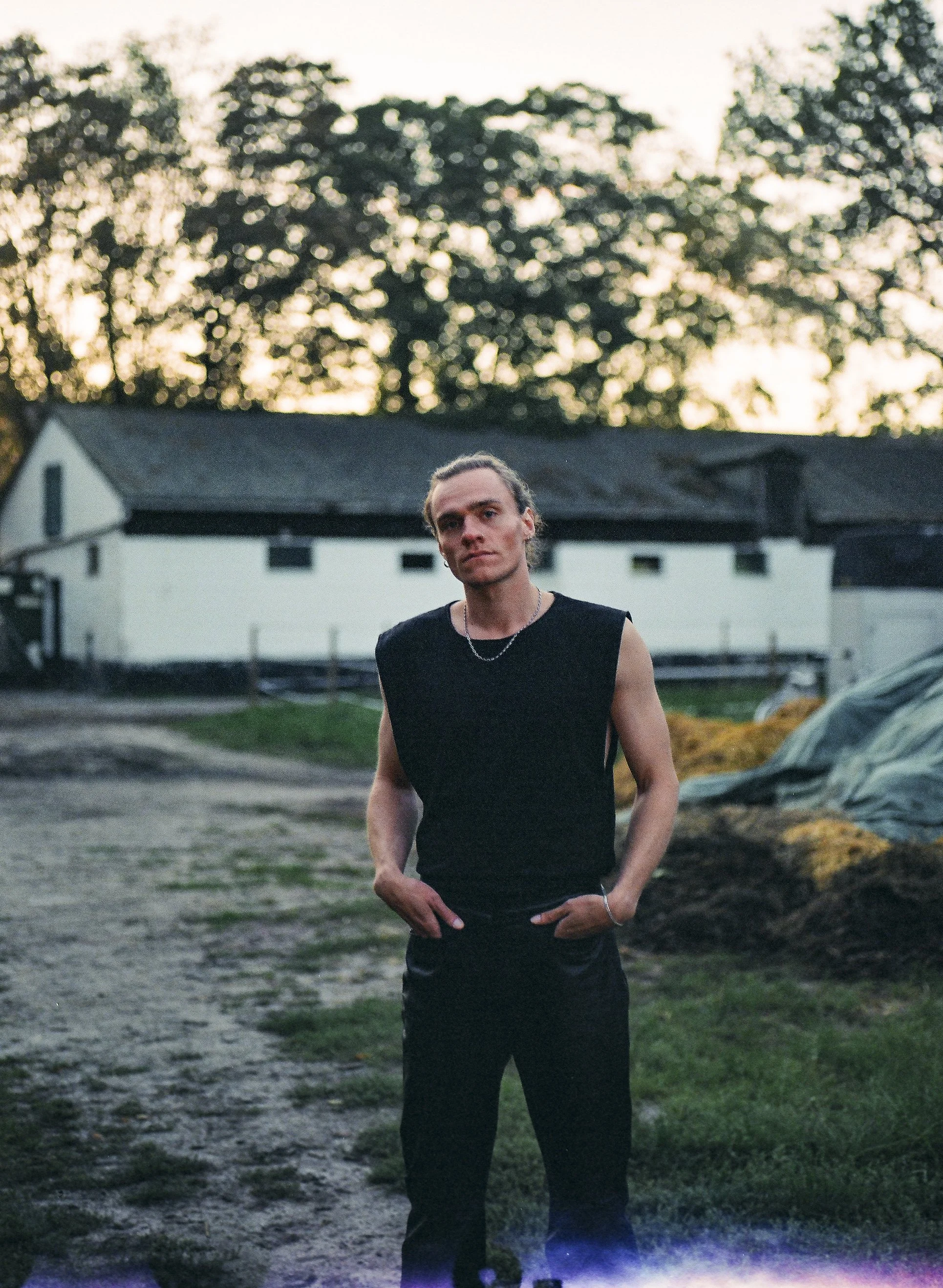 Junger Mann in schwarzem Hemd steht im Freien bei Sonnenuntergang, im Hintergrund Gebäuden und Bäume.