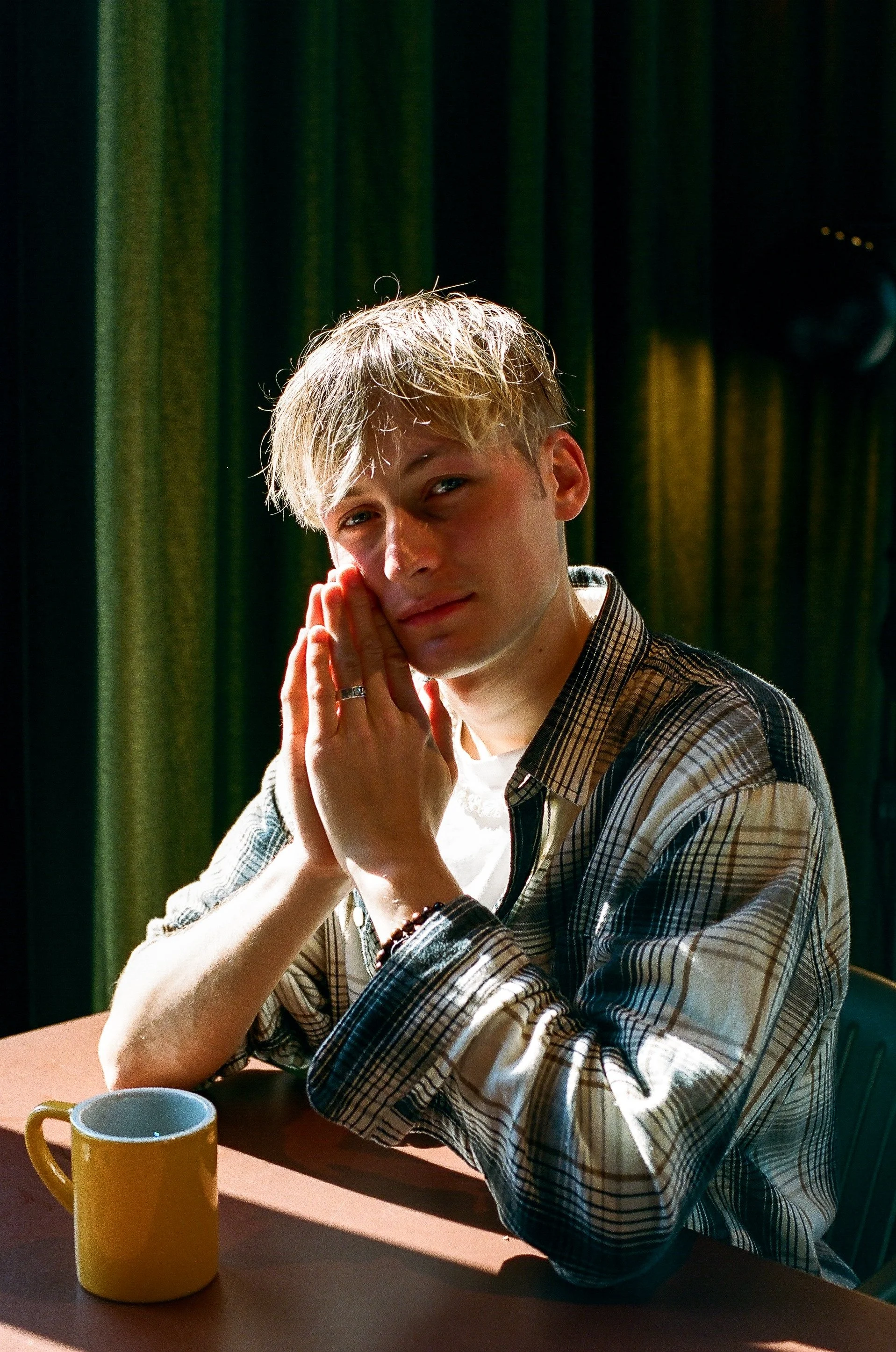 Ein junger Mann mit blondem Haar sitzt an einem Tisch, hält seine Hände an sein Gesicht und schaut in die Kamera. Es gibt eine gelbe Kaffeetasse vor ihm und grüne Vorhänge im Hintergrund.