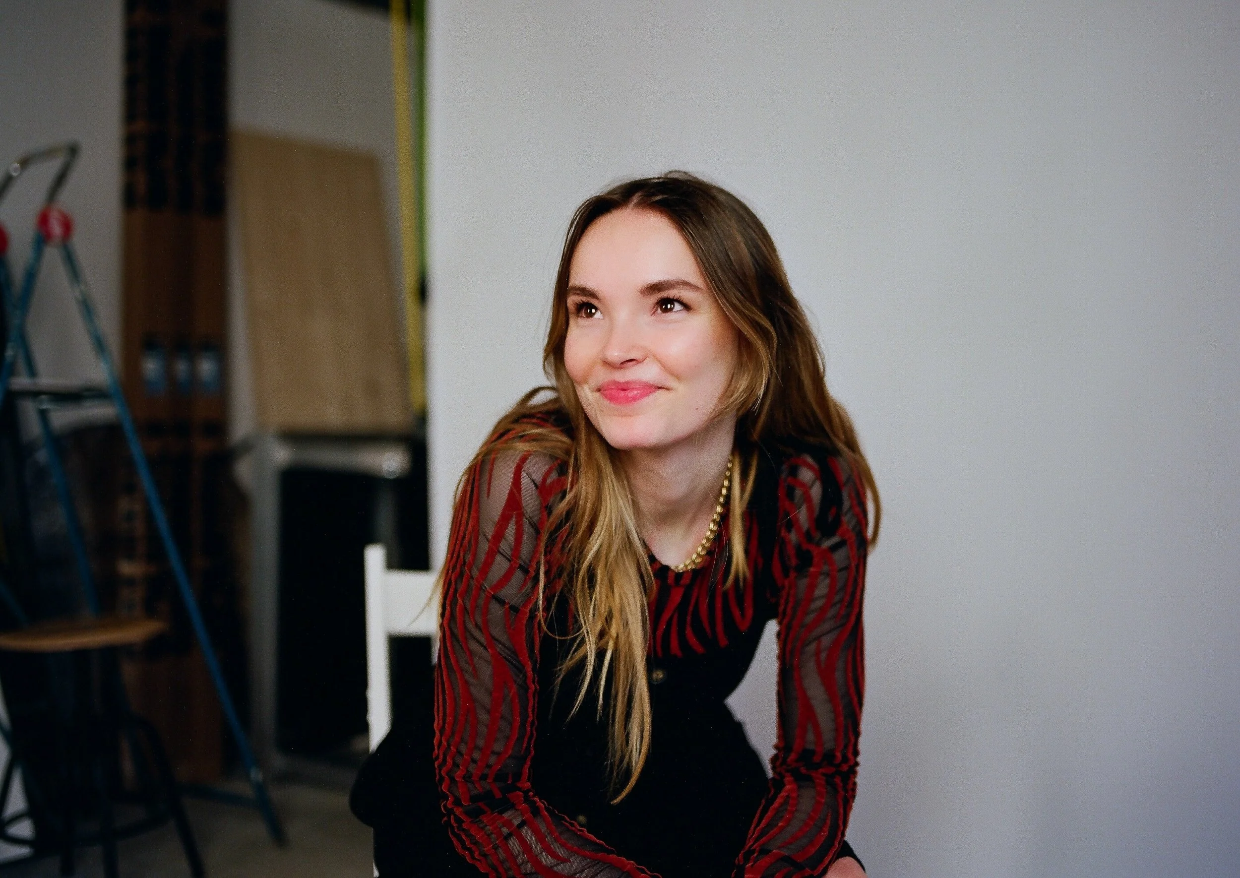 Junge Frau mit langem, gewelltem Haar, sitzt vor einer neutralen Wand, trägt eine schwarze Jacke mit roten Streifen, lächelt und schaut leicht nach oben.