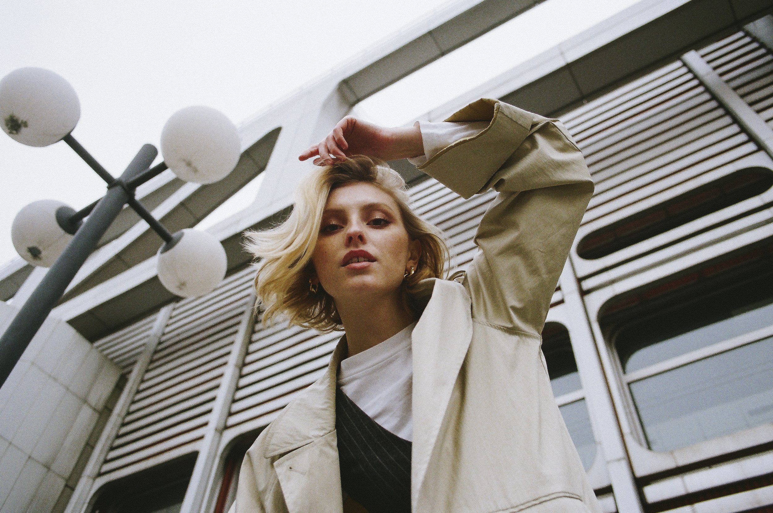 Junge Frau mit blonden Haaren posiert vor einem modernen Gebäude mit großen Fenstern und vertikalen Lamellen, trägt beige Jacke und weiße T-Shirt, und liegt die Hand auf dem Kopf.