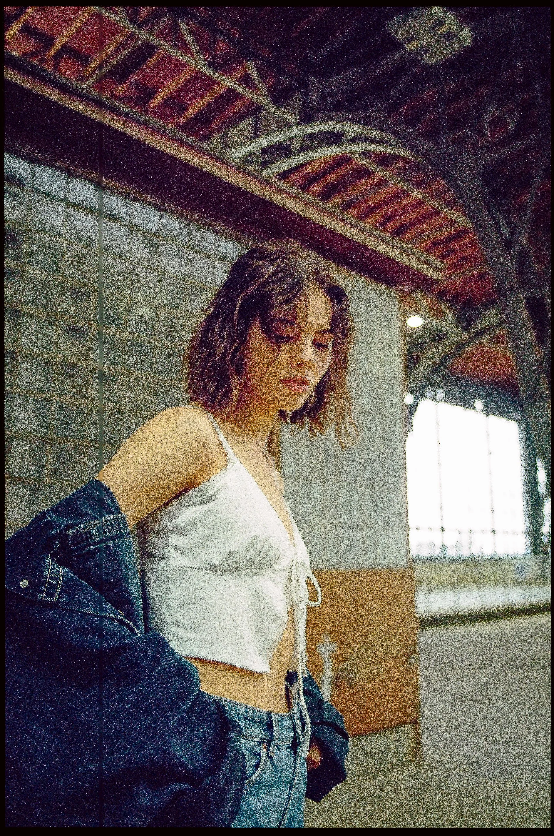 Junge Frau steht in einem Innenraum mit großen Fenstern im Hintergrund, trägt ein weißes Top und eine offene Jeansjacke, wirkt nachdenklich.