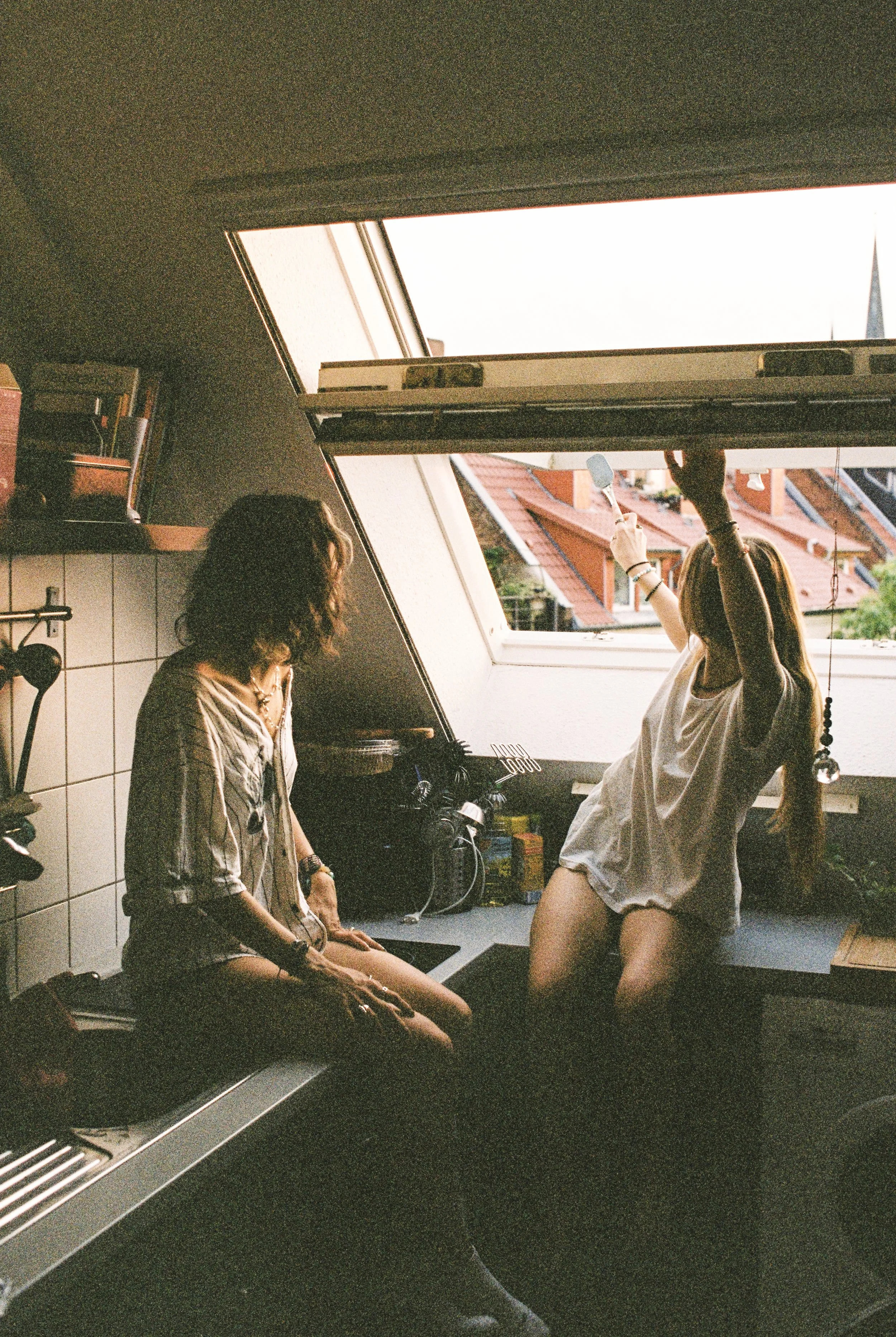 Zwei Frauen im Küchenraum eines Dachgeschosses, eine sitzt auf der Küchentheke, die andere ist am Fenster im Stehen und öffnet es, bei Sonnenlicht.