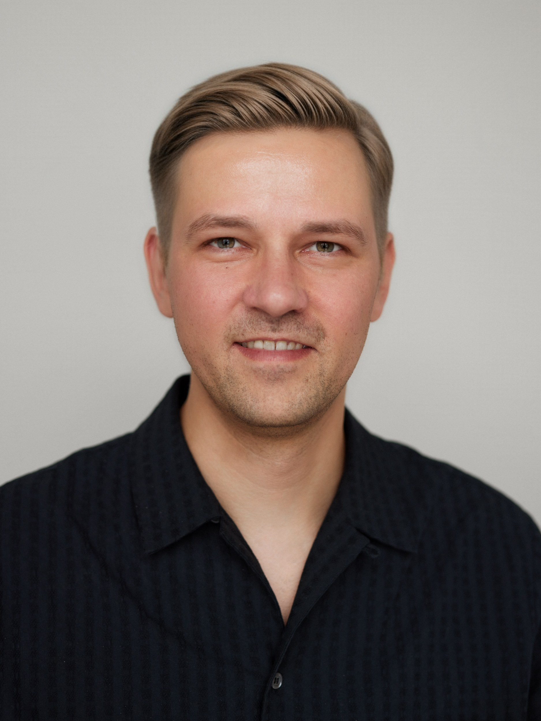 Close-up of a young man with blonde hair, blue eyes, light skin, and a subtle smile, wearing a black textured button-up shirt against a plain light gray background.