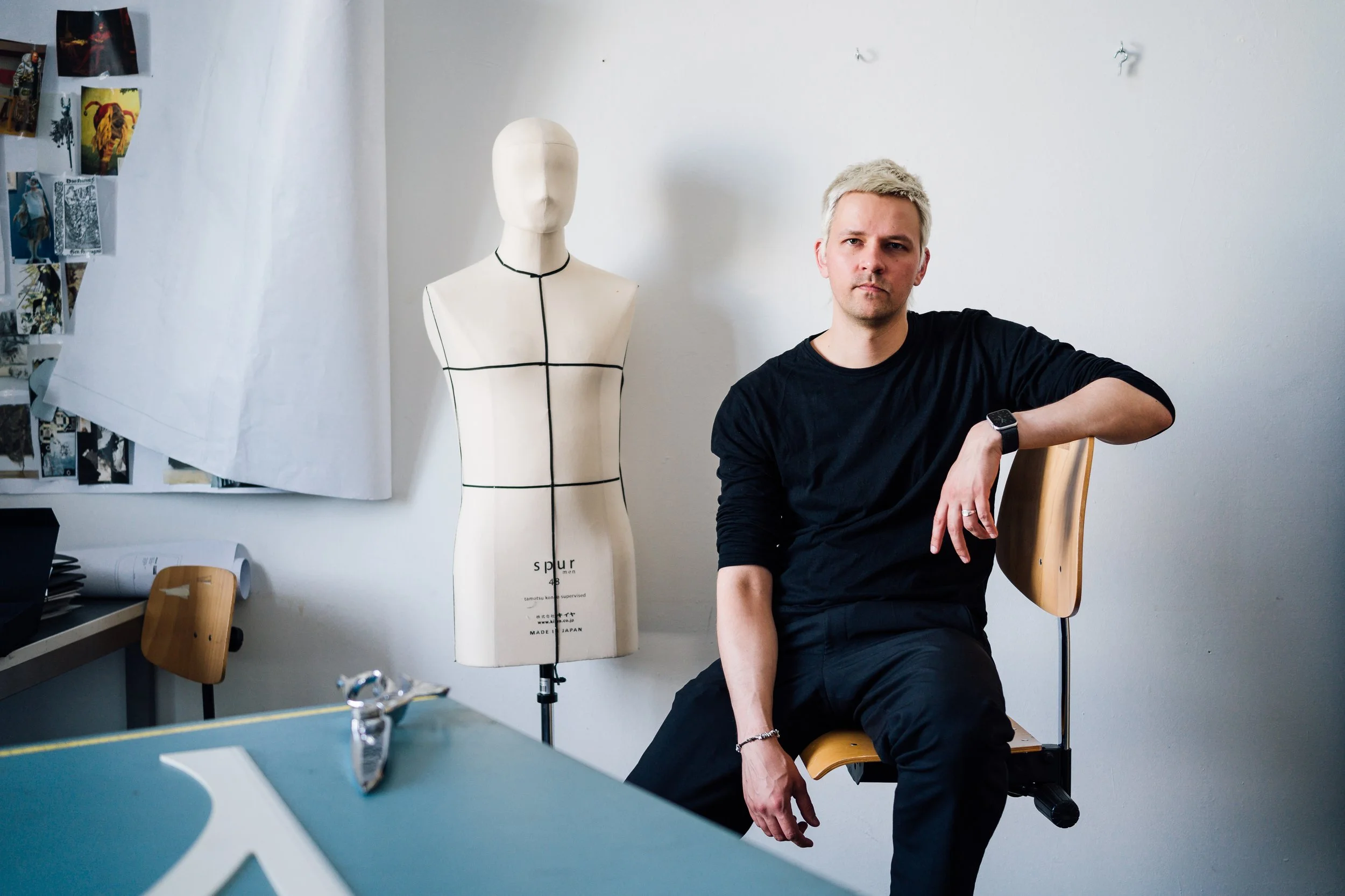 A young man with platinum blond hair sitting on a chair in a room with white walls. Behind him is a mannequin with black lines marking design sections, labeled 'spur', made in Japan. To the left, there are photo collages and a desk with papers and a wooden chair.