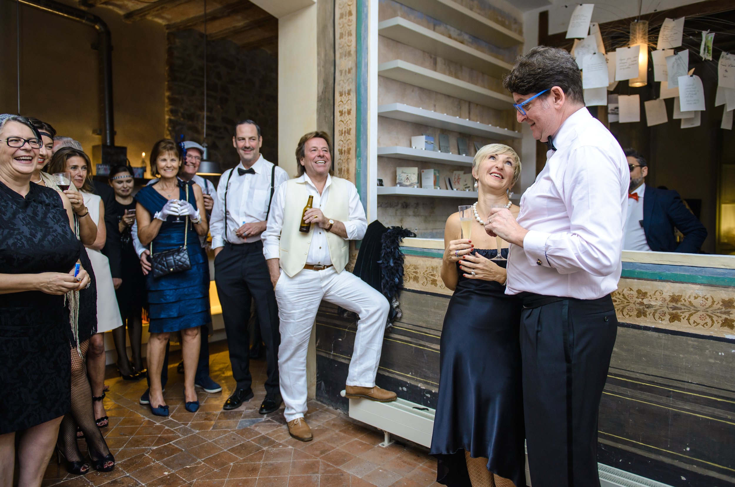 Grupo de personas elegantemente vestidas en una fiesta dentro de un lugar con decoración clásica, algunas están sonriendo y conversando, y otras sostienen bebidas.