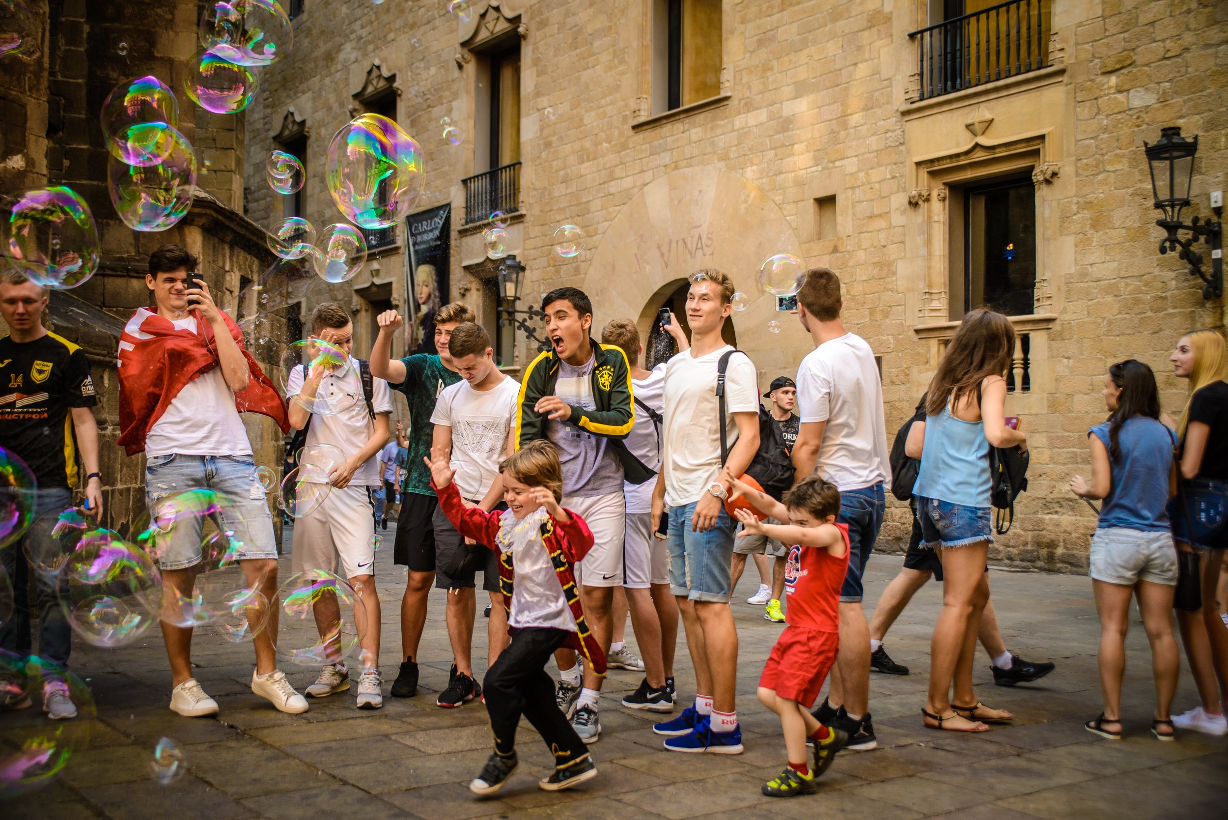 Grupo de personas, incluyendo niños, interactuando con burbujas en una calle de una ciudad con edificios antiguos de piedra. Todos parecen estar disfrutando del momento.