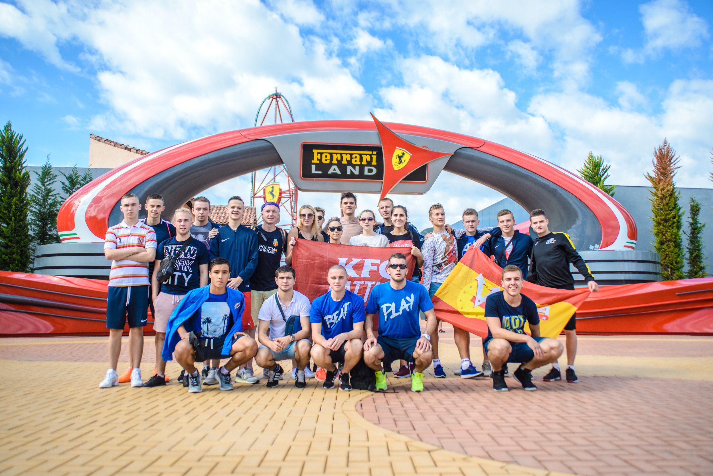 Grupo de personas frente al cartel de Ferrari Land, con algunas sosteniendo banderas. Cielo despejado y árboles en el fondo.