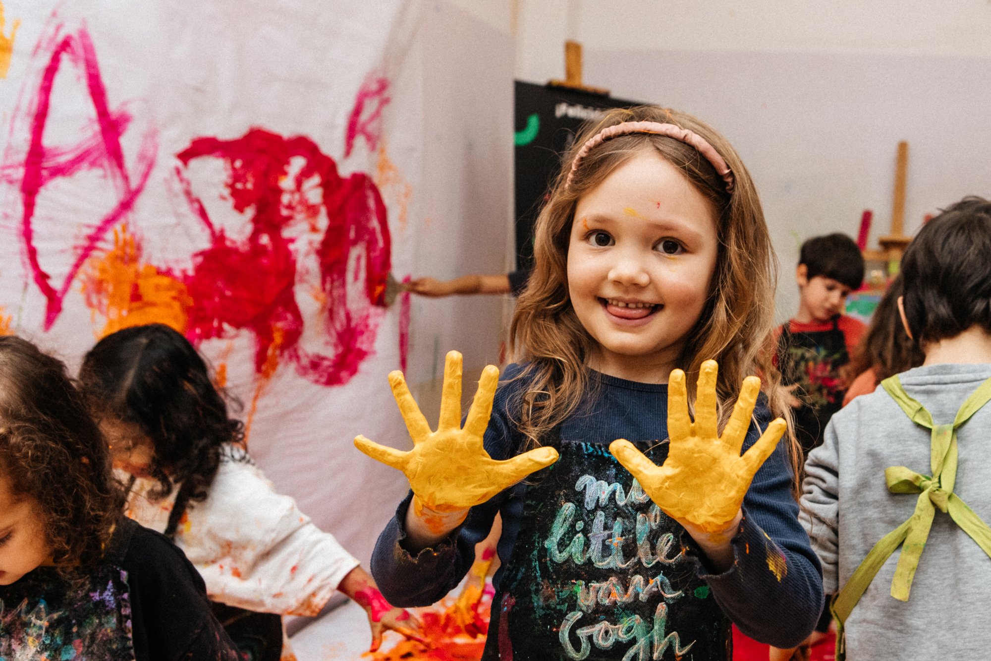 Niños pintando en una clase de arte, una niña muestra sus manos cubiertas de pintura amarilla sonriente.