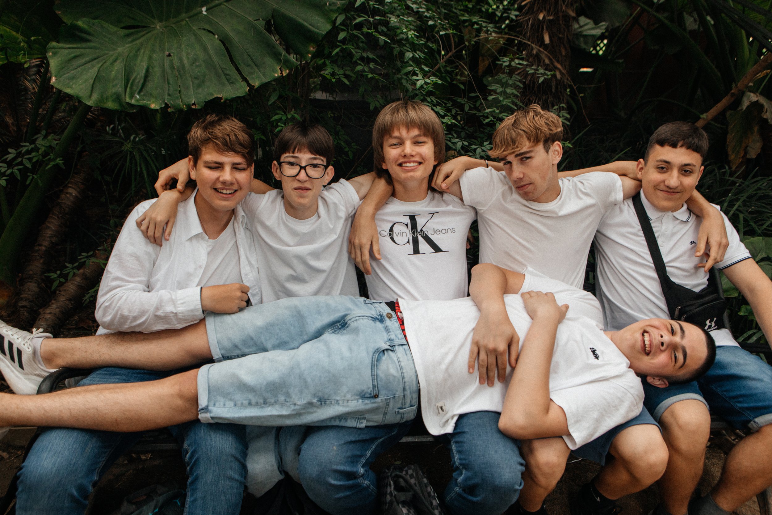 Grupo de jóvenes abrazados, vistiendo camisetas blancas, en un entorno de jardín con hojas grandes.