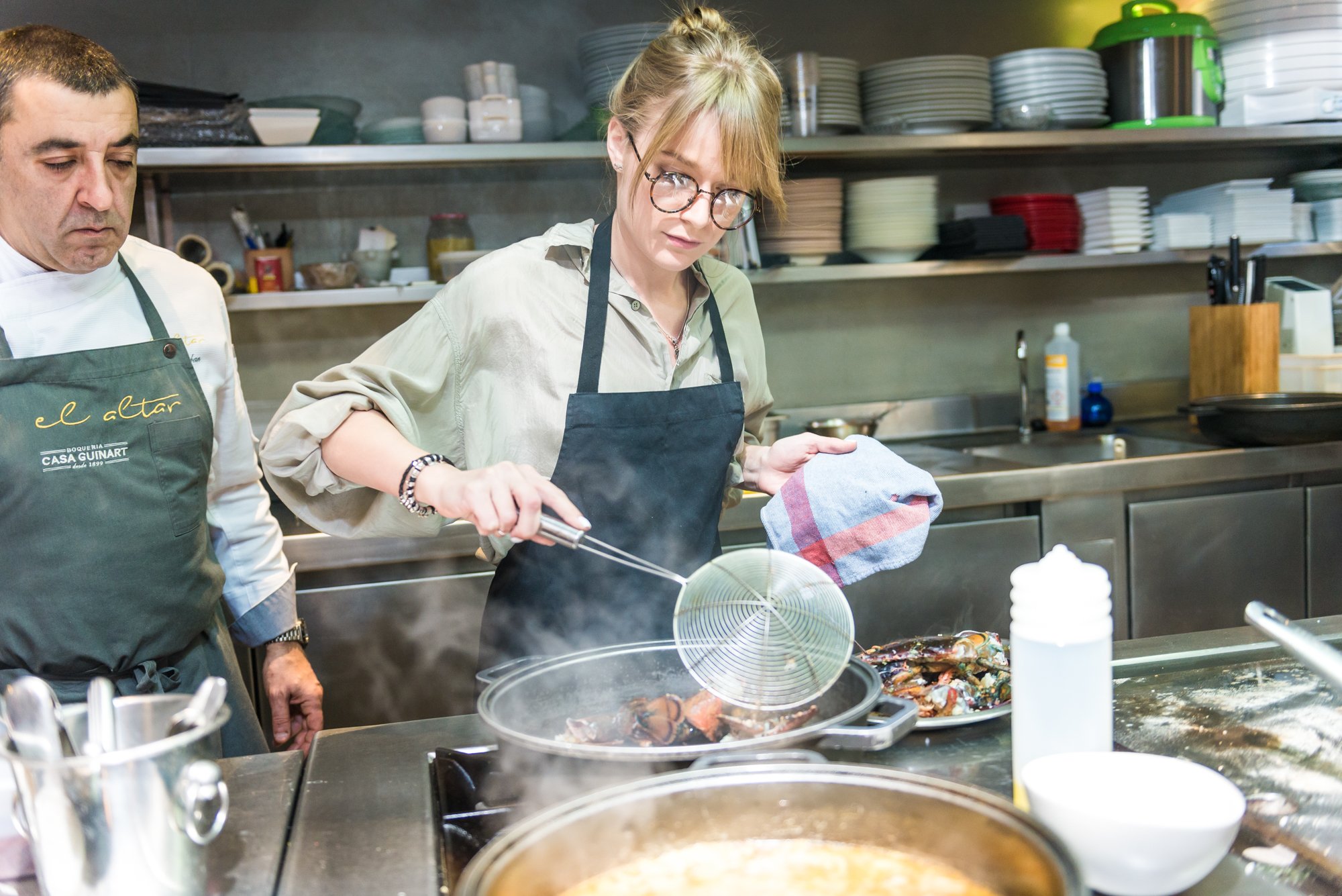 Dos personas cocinando en una cocina profesional, una mujer usando un delantal y moviendo alimentos en una sartén mientras un hombre la observa. Hay utensilios y platos en los estantes del fondo.