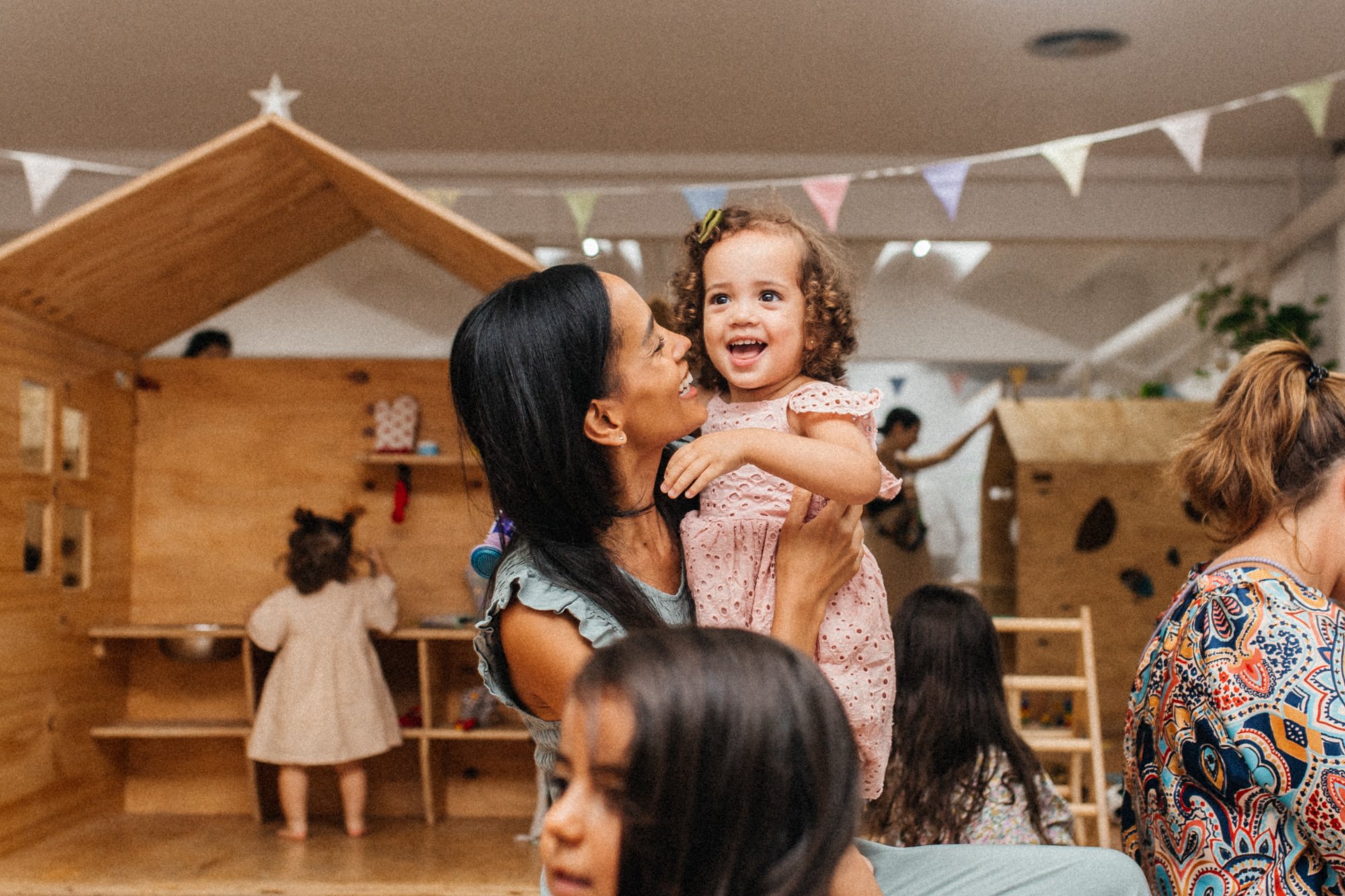 Una mujer sostiene a una niña sonriente en un ambiente interior con decoración infantil y otras personas alrededor.