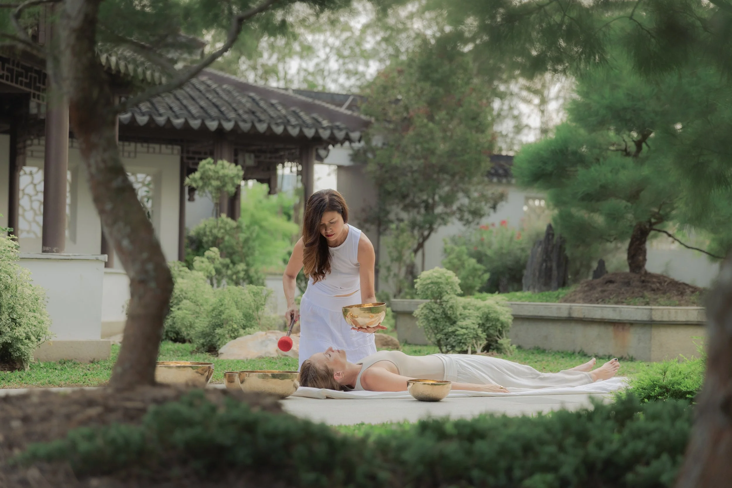 A woman dressed in white is lying down on a mat in a garden, receiving an energy healing treatment from a practitioner who is sprinkling water from a bowl onto her forehead, with traditional Asian architecture and lush greenery in the background.