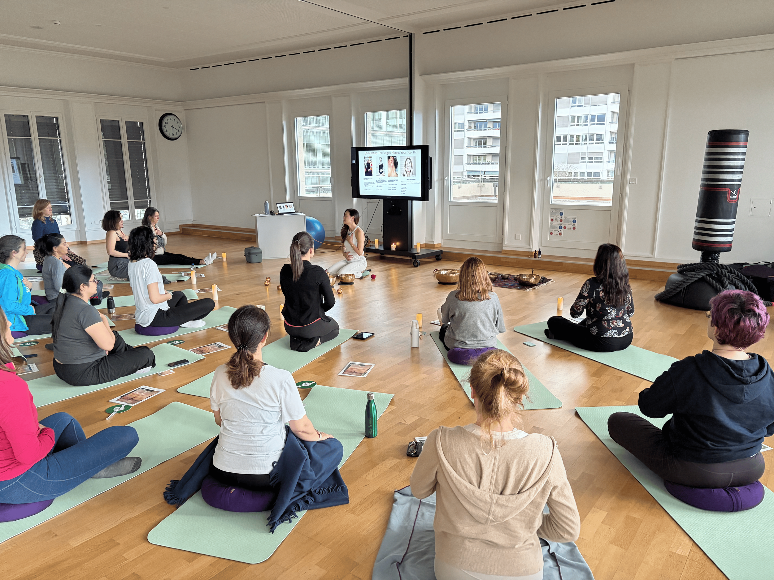 A group of people practicing yoga or meditation in a spacious room with large windows, sitting on yoga mats in a circle around a woman who appears to be leading the session, with a large screen displaying yoga or health-related information.