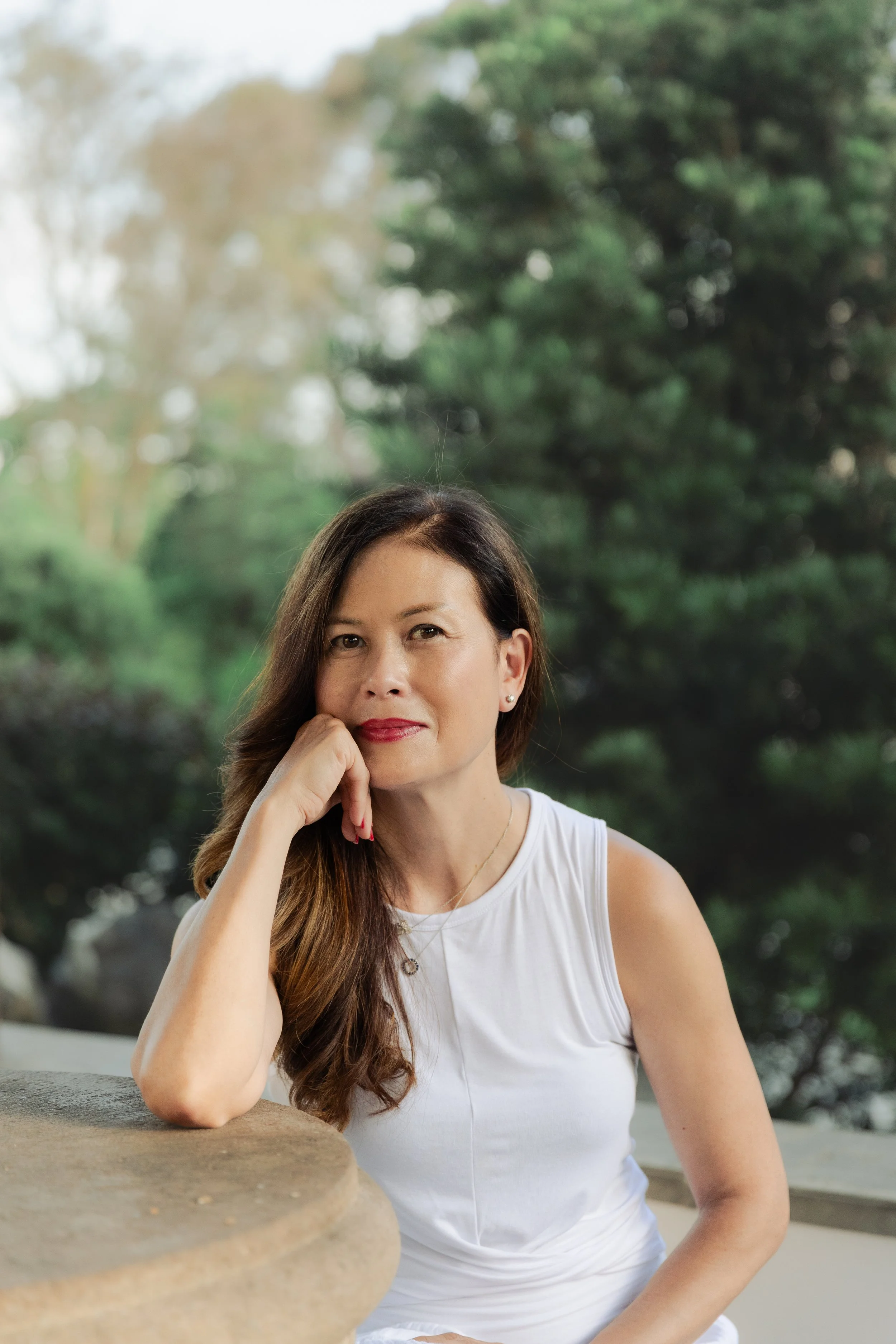 A woman with long brown hair sitting outdoors near a stone bench, resting her chin on her hand, with trees and cloudy sky in the background.