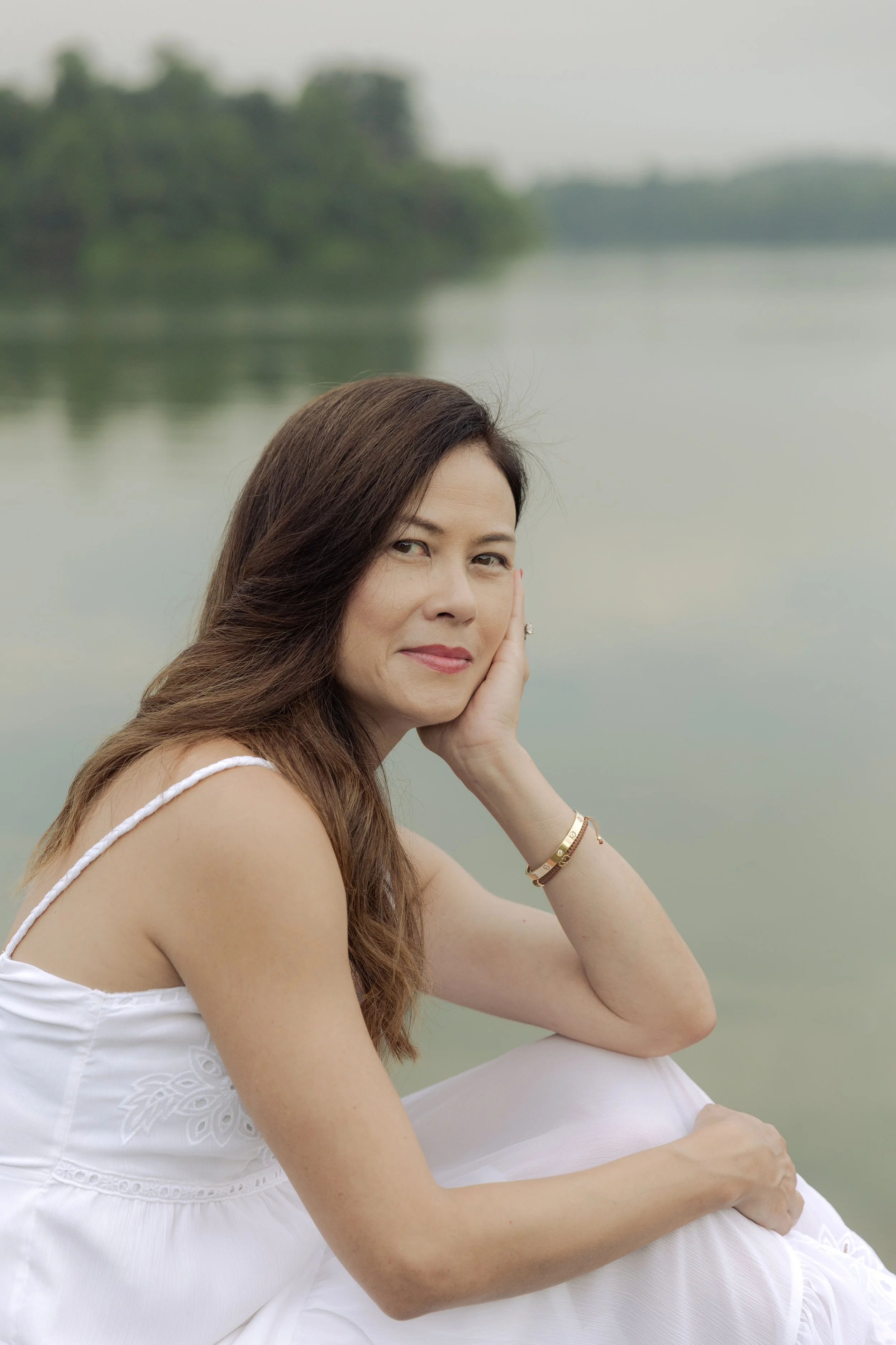 A woman with long brown hair, sitting outdoors by a body of water with trees in the background, wearing a white dress and jewelry, resting her head on her hand.