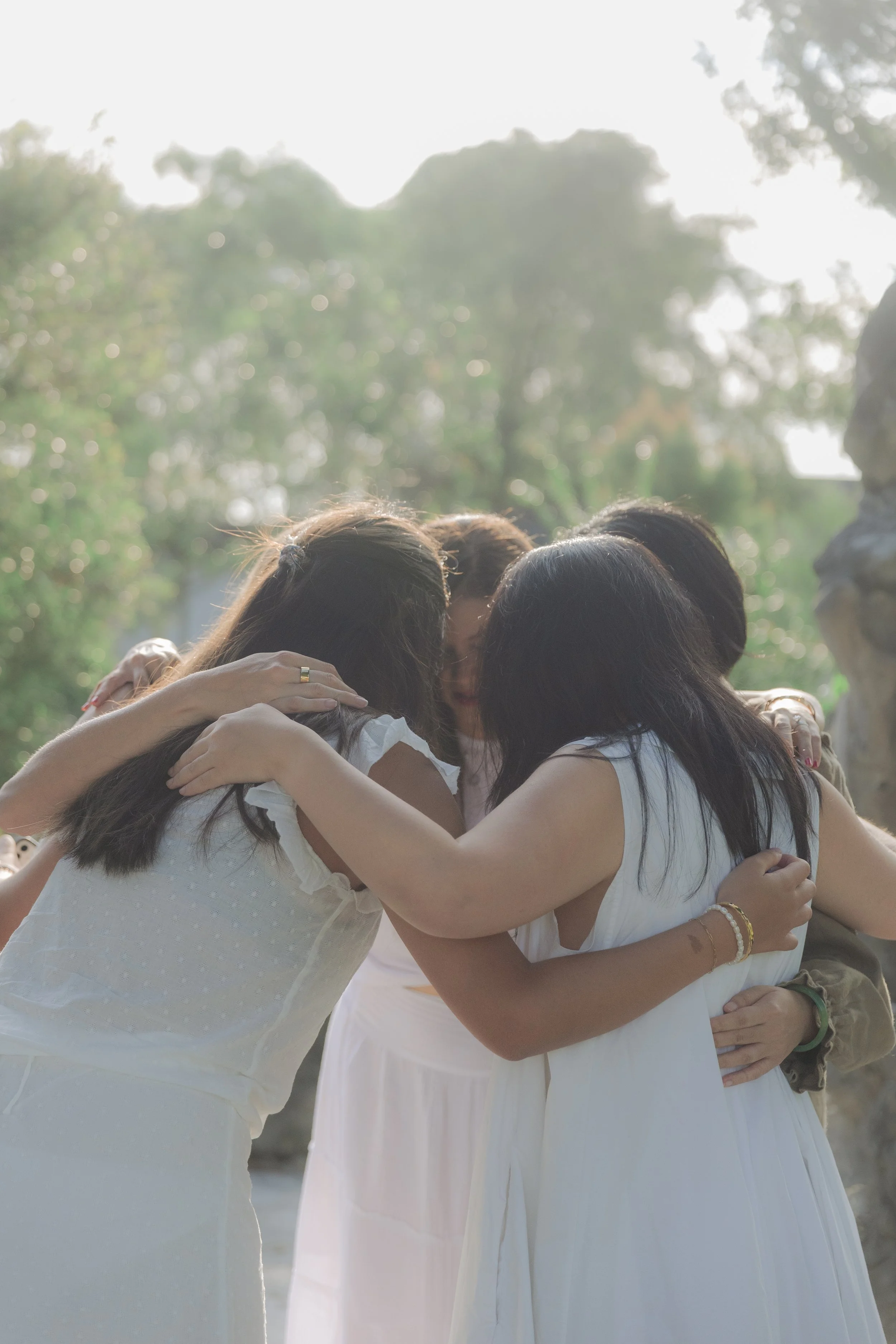 Group of women hugging each other outdoors, all wearing white dresses, with sunlight and trees in the background.