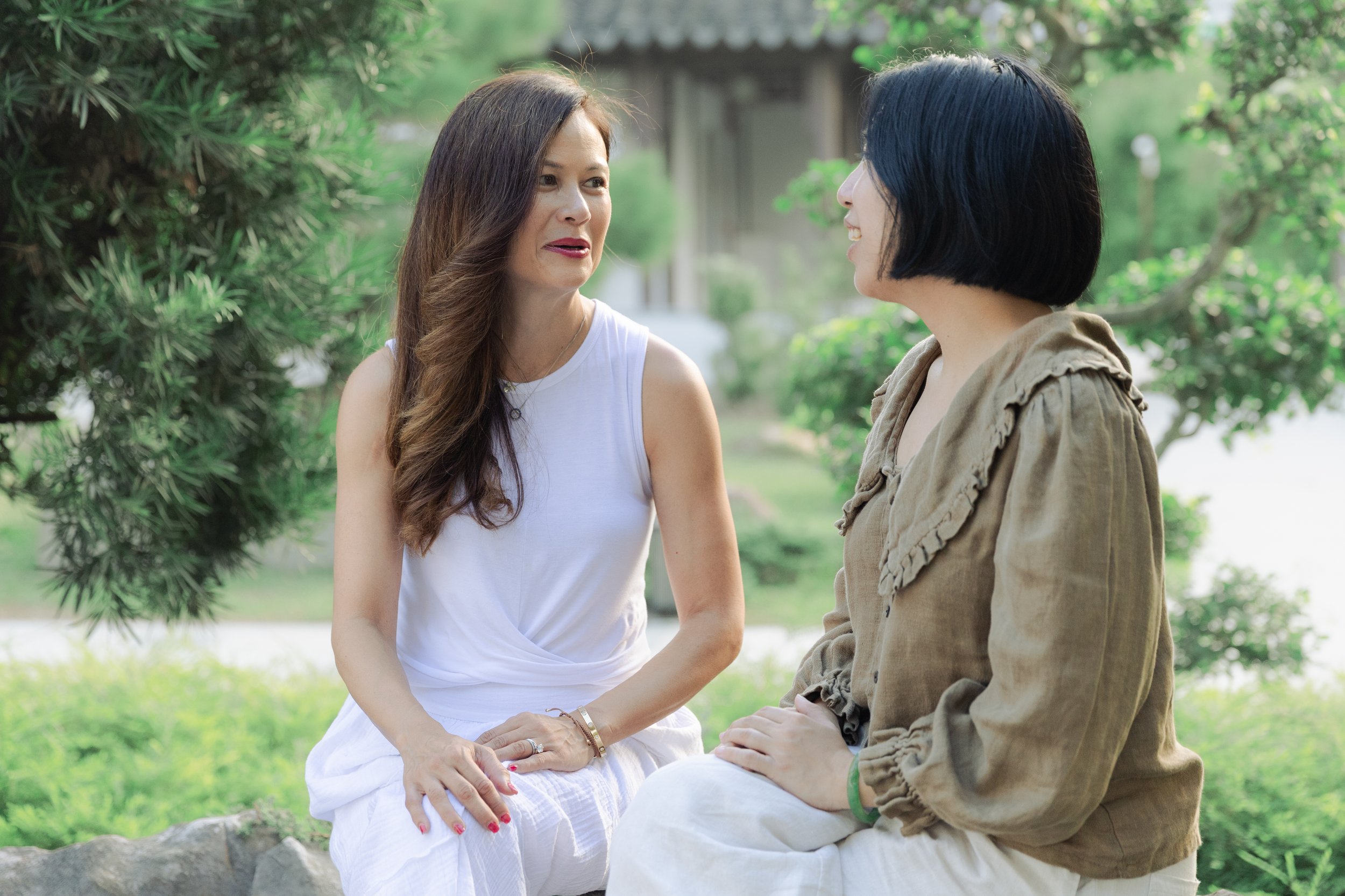 Two women sitting outdoors in a park, engaged in a conversation. One woman has long brown hair, wearing a white sleeveless top, and the other woman has short black hair, wearing a beige jacket.