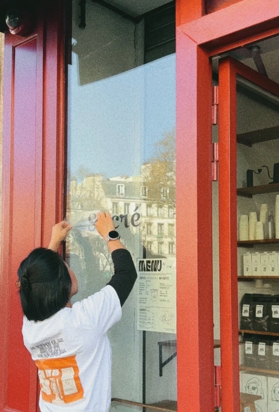 Une personne colle un sticker sur une vitre d'une boutique de café. La vitrine a un cadre rouge, et il y a un menu affiché à l'intérieur. Des bâtiments résidentiels sont visibles en arrière-plan.