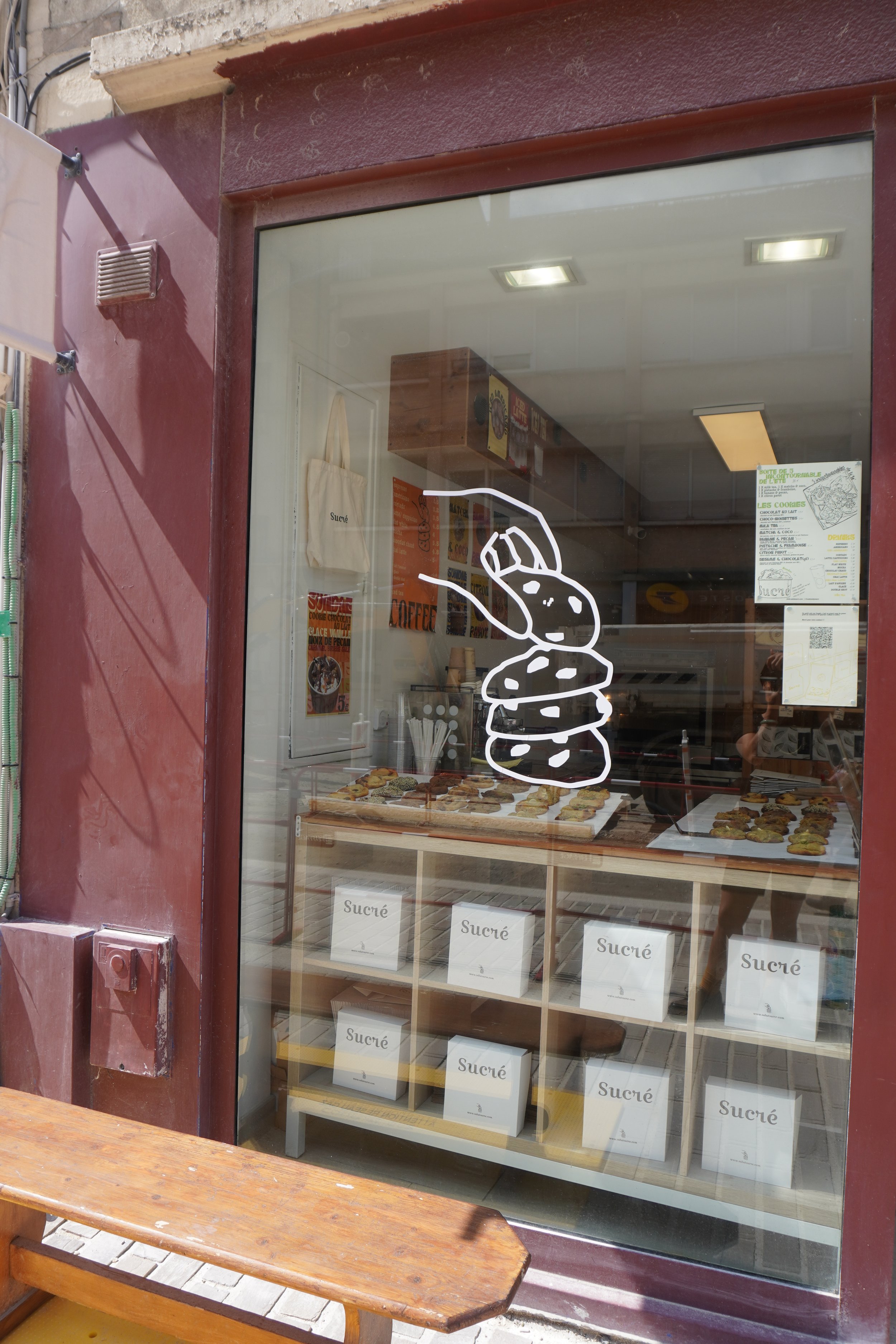 Vitrine d'une boutique avec une illustration d'une queue de bonbons blanc. À l'intérieur, il y a des boîtes de sucre avec le mot 'Sucré' écrit dessus et un comptoir avec des biscuits ou des pâtisseries.