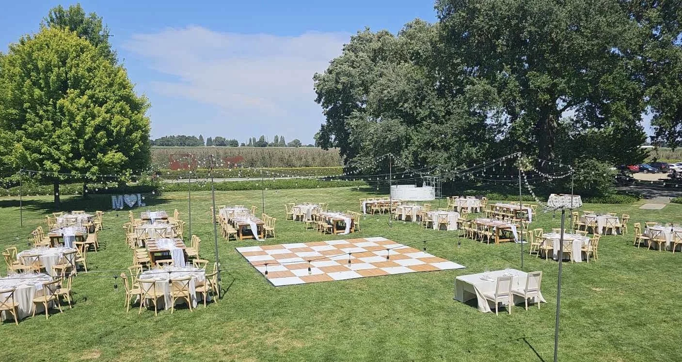 Outdoor event setup with multiple round tables with white tablecloths and chairs, a large checkerboard dance floor in the center, string lights overhead, and large trees in the background on a grassy lawn.