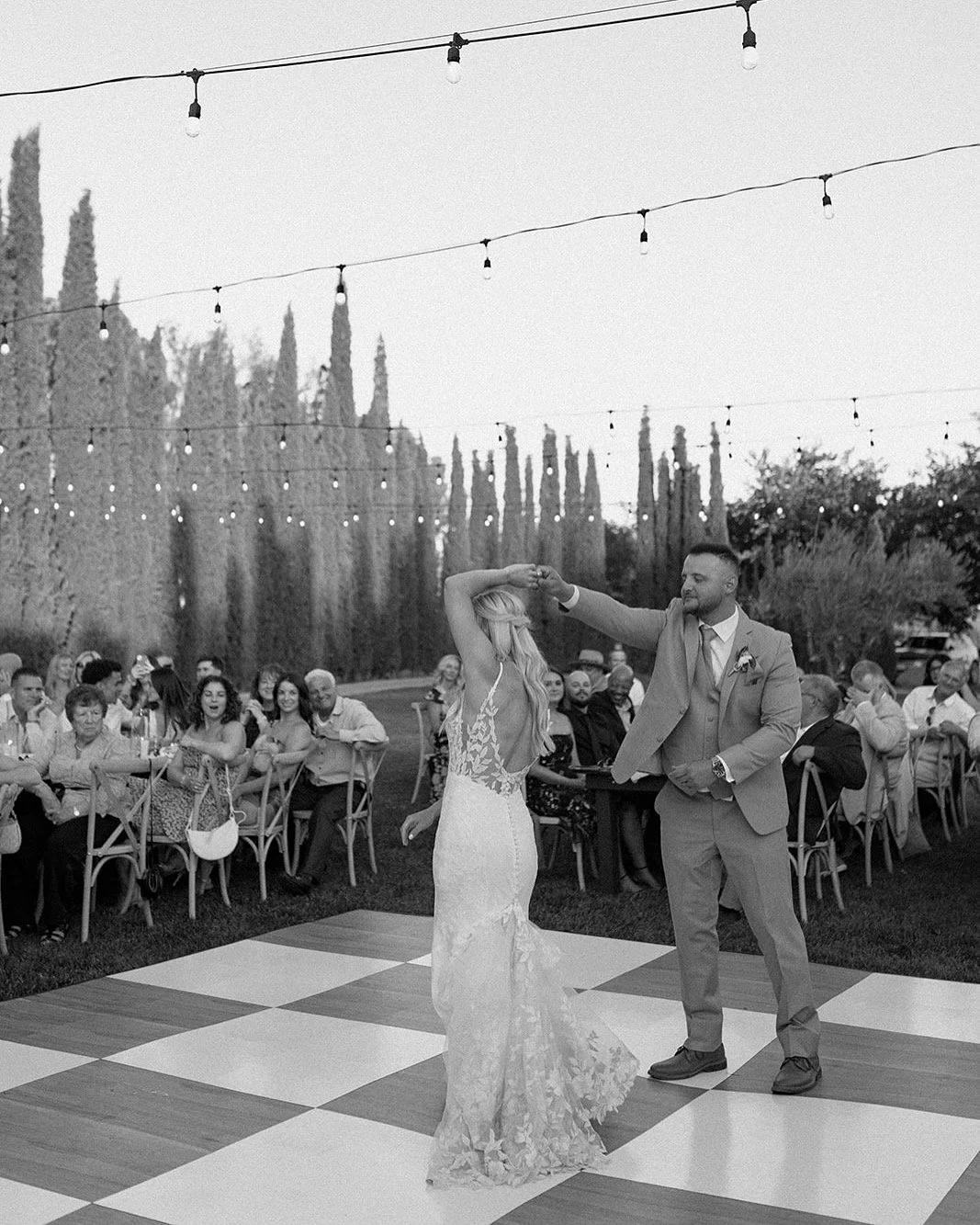 A black and white photo of a wedding dance outdoors with guests seated at tables in the background, string lights overhead, a bride in a lace gown dancing with a groom in a suit.