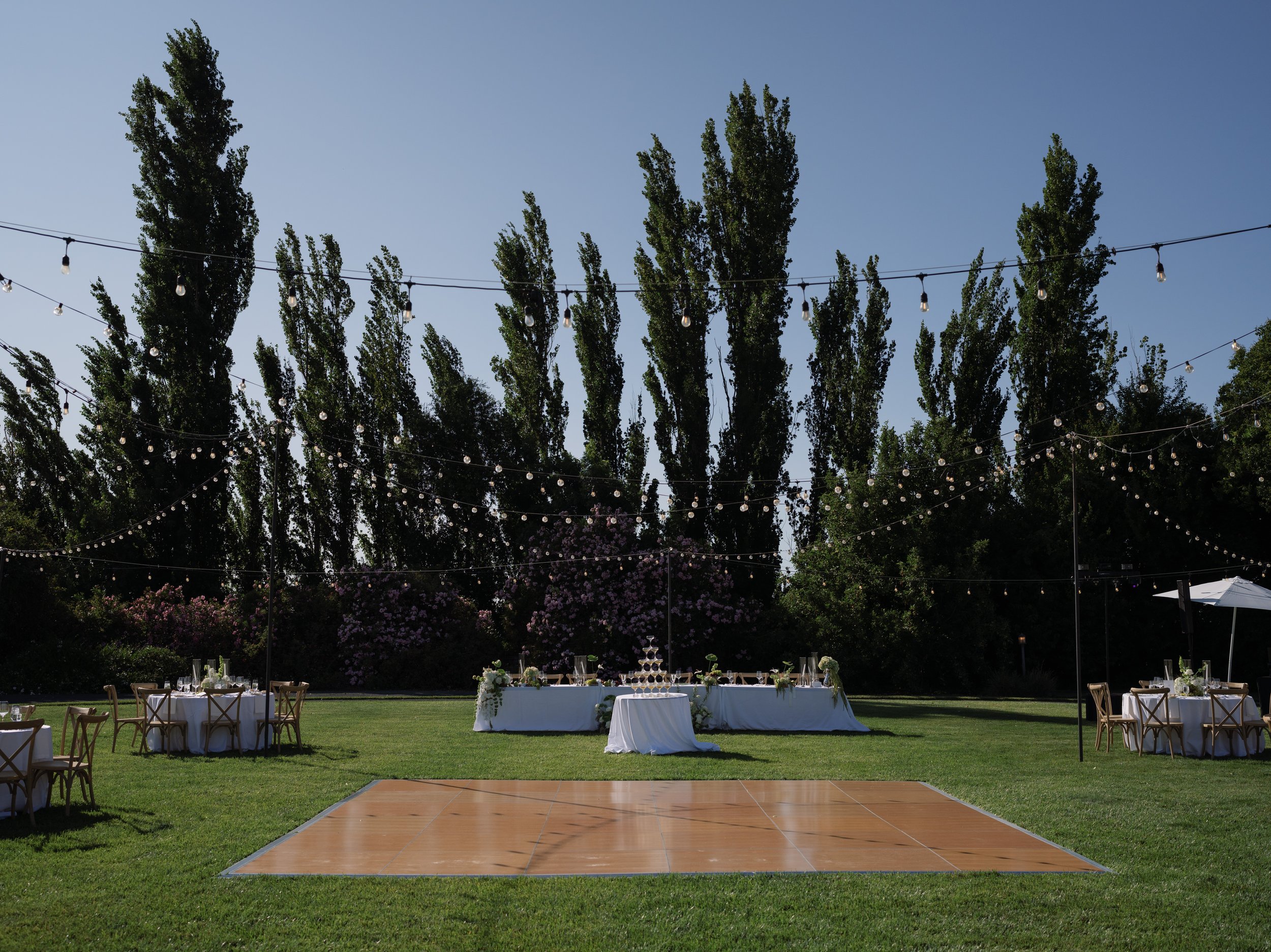 Outdoor wedding reception setup with round tables, white tablecloths, wooden chairs, string lights overhead, a dance floor in the foreground, and a long head table in the background on a grassy lawn.