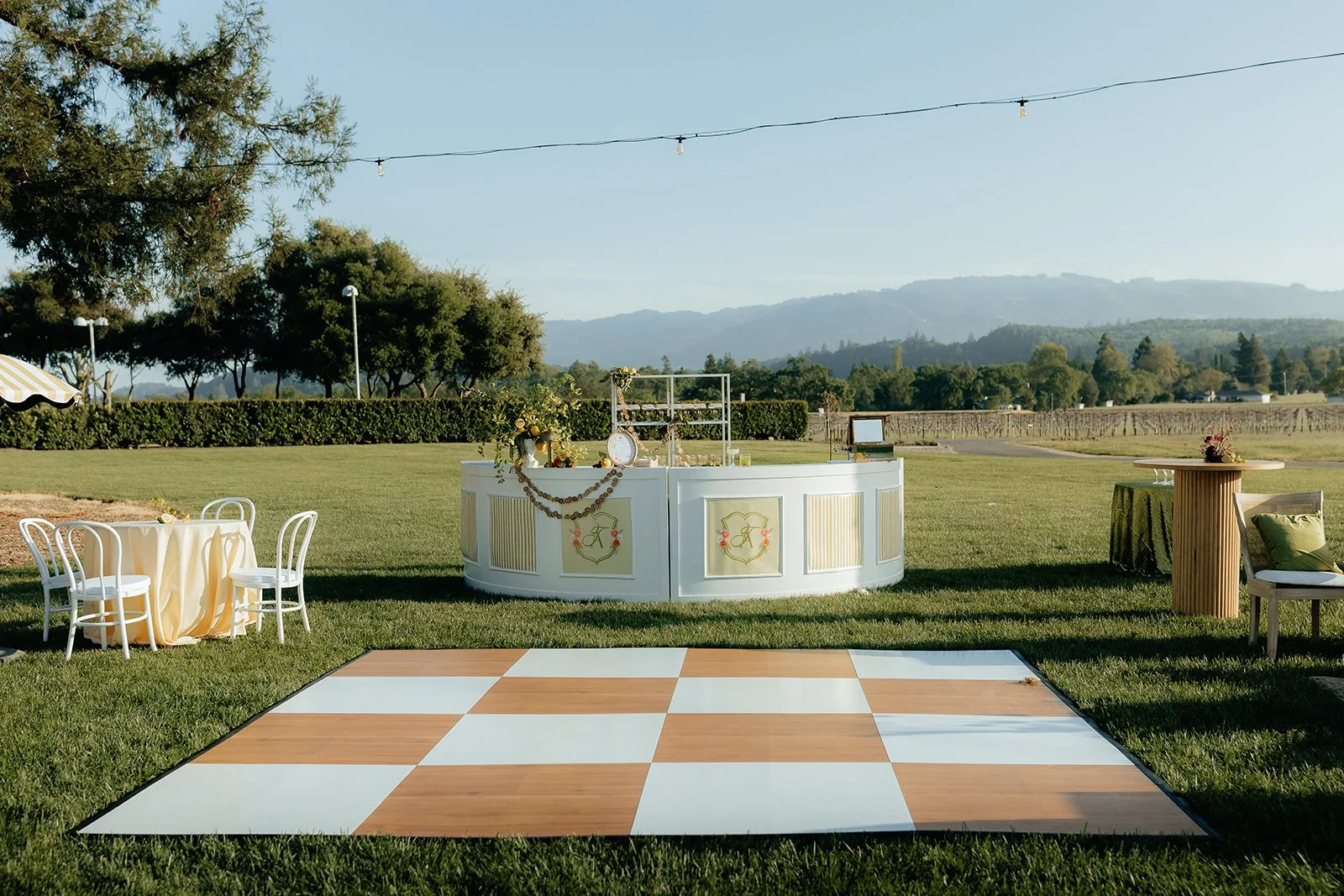 Outdoor event setup on a grassy field with a white bar, a dance floor with a checkered pattern, a round table with white chairs, and a small high-top table, surrounded by trees and mountains in the background under a clear sky.