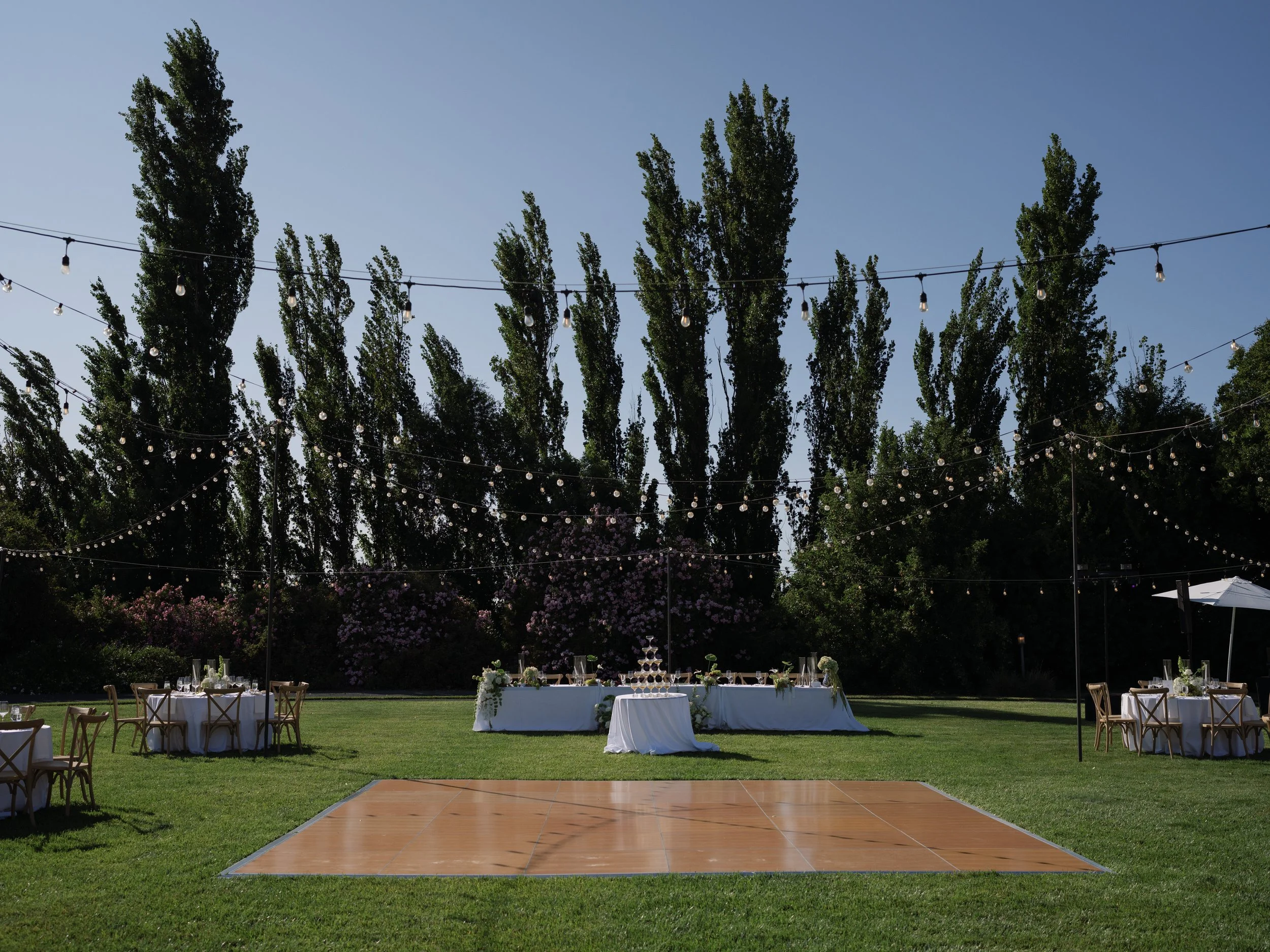 Outdoor wedding reception setup with tables, chairs, and string lights on a grassy lawn, with a dance floor in the foreground and trees in the background under a clear sky.
