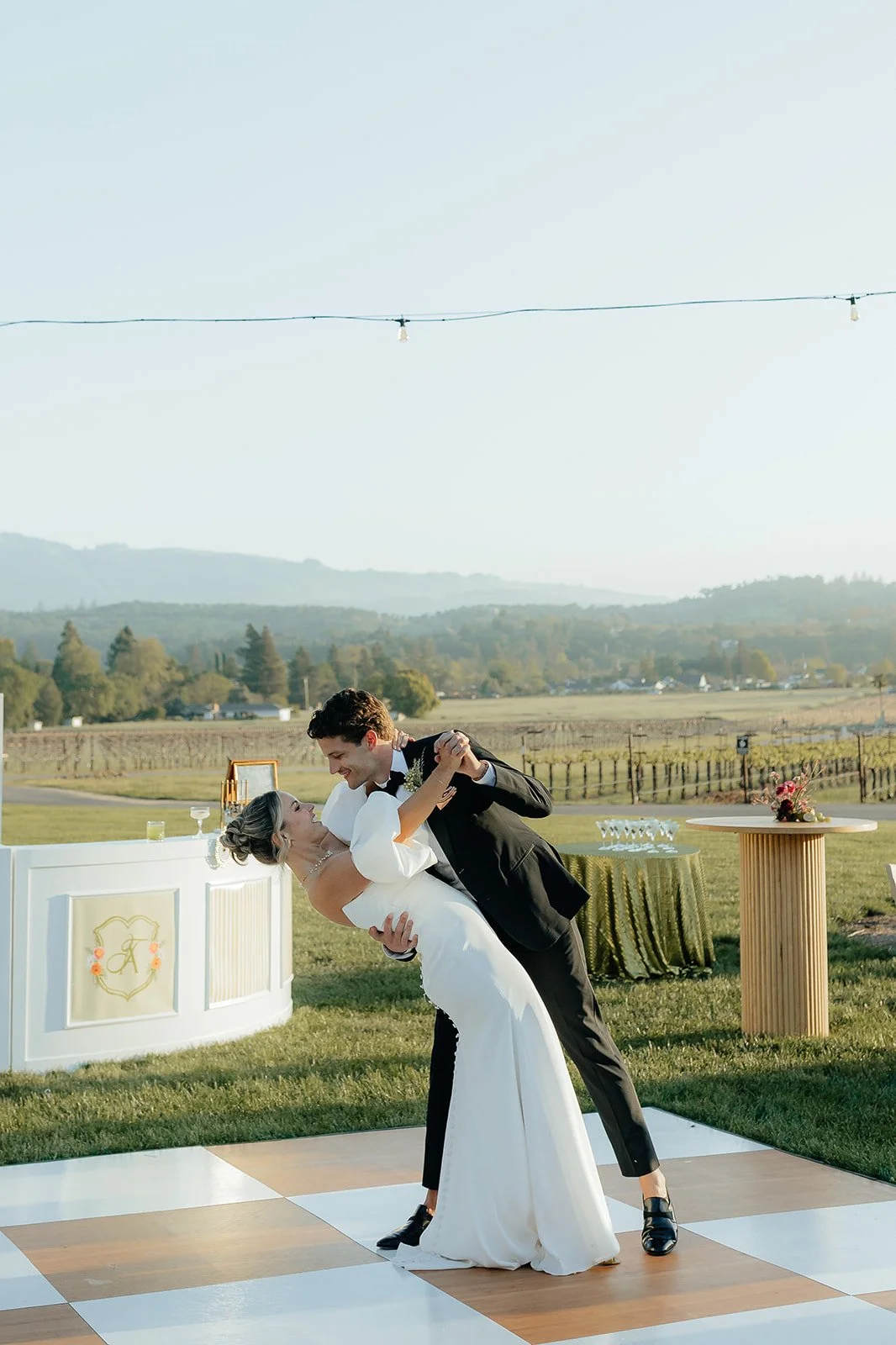 A bride and groom dancing outdoors at their wedding reception on a checkered dance floor, with a scenic vineyard and rolling hills in the background, under string lights.