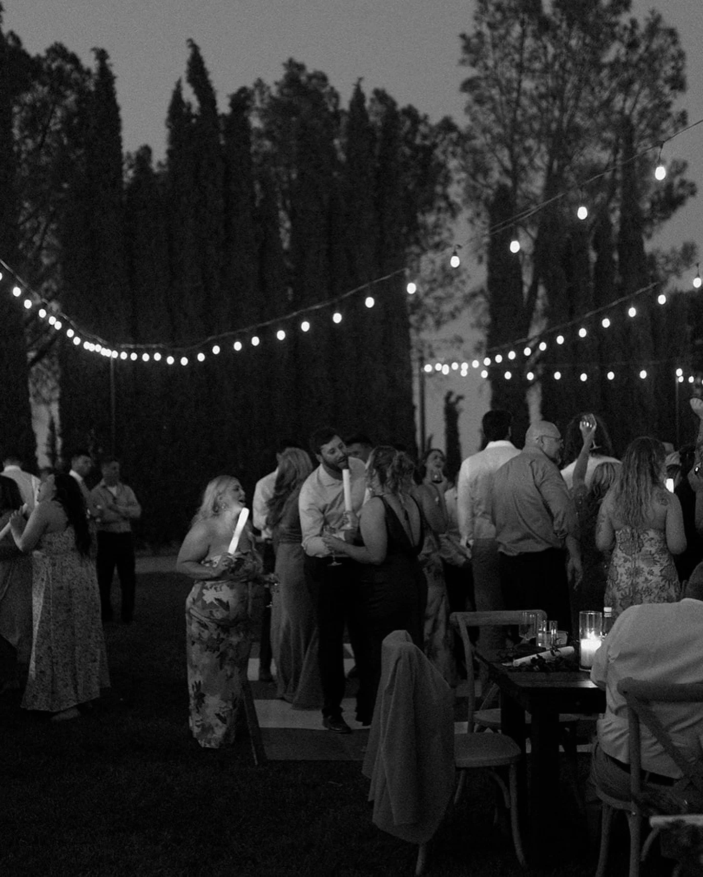 People dancing and socializing at an outdoor evening event under string lights, with trees in the background.