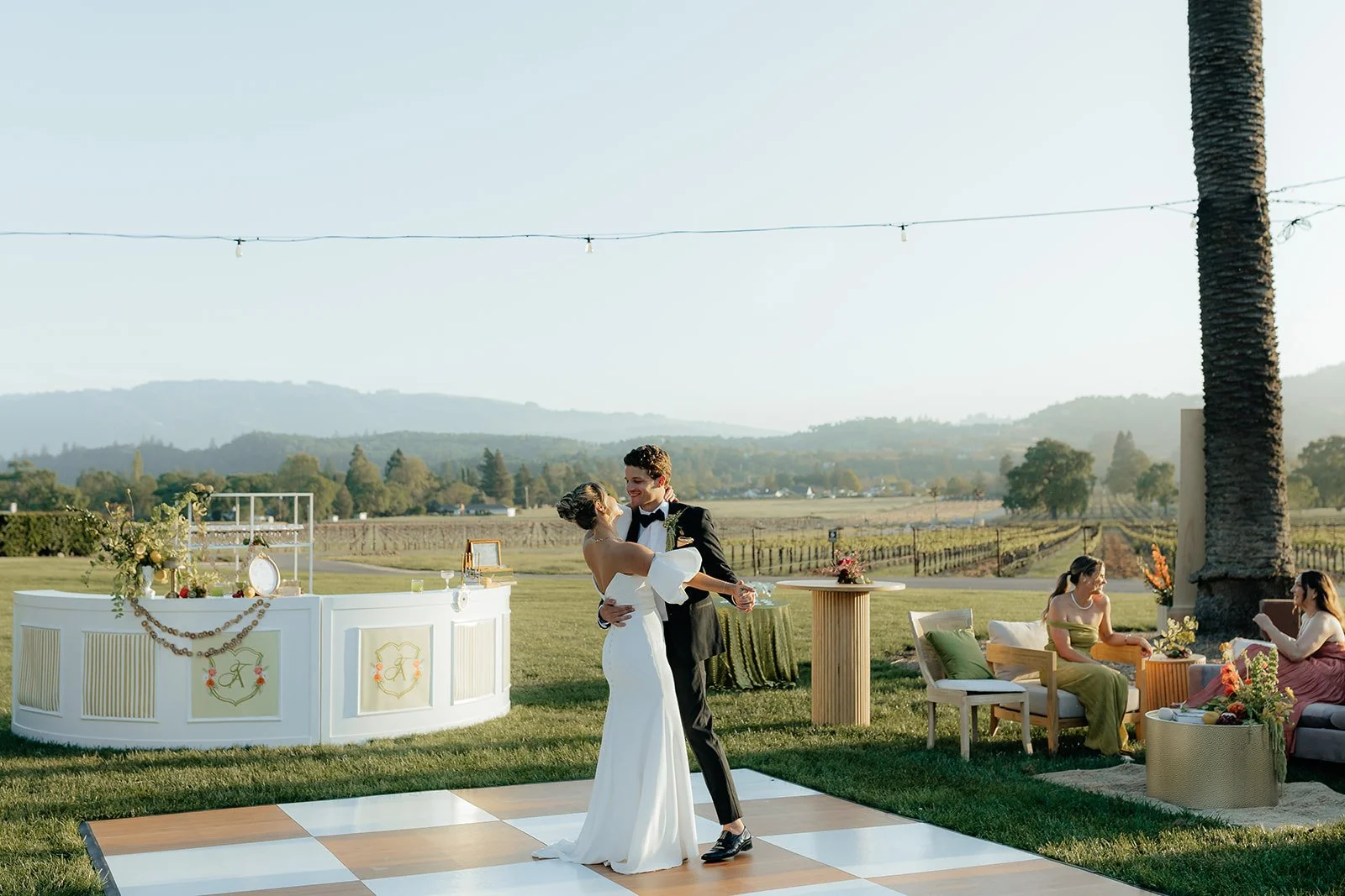 A bride and groom dancing outdoors at a wedding reception in a scenic vineyard with rolling hills in the background, with two women sitting on a couch nearby.