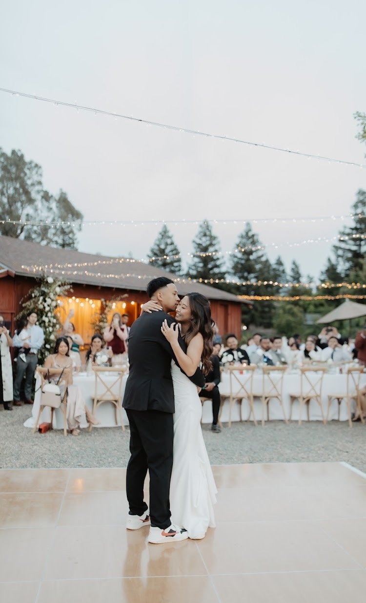 A bride and groom dancing during their outdoor wedding reception at sunset, with guests seated at tables and celebrating in the background, string lights overhead, and trees surrounding the venue.