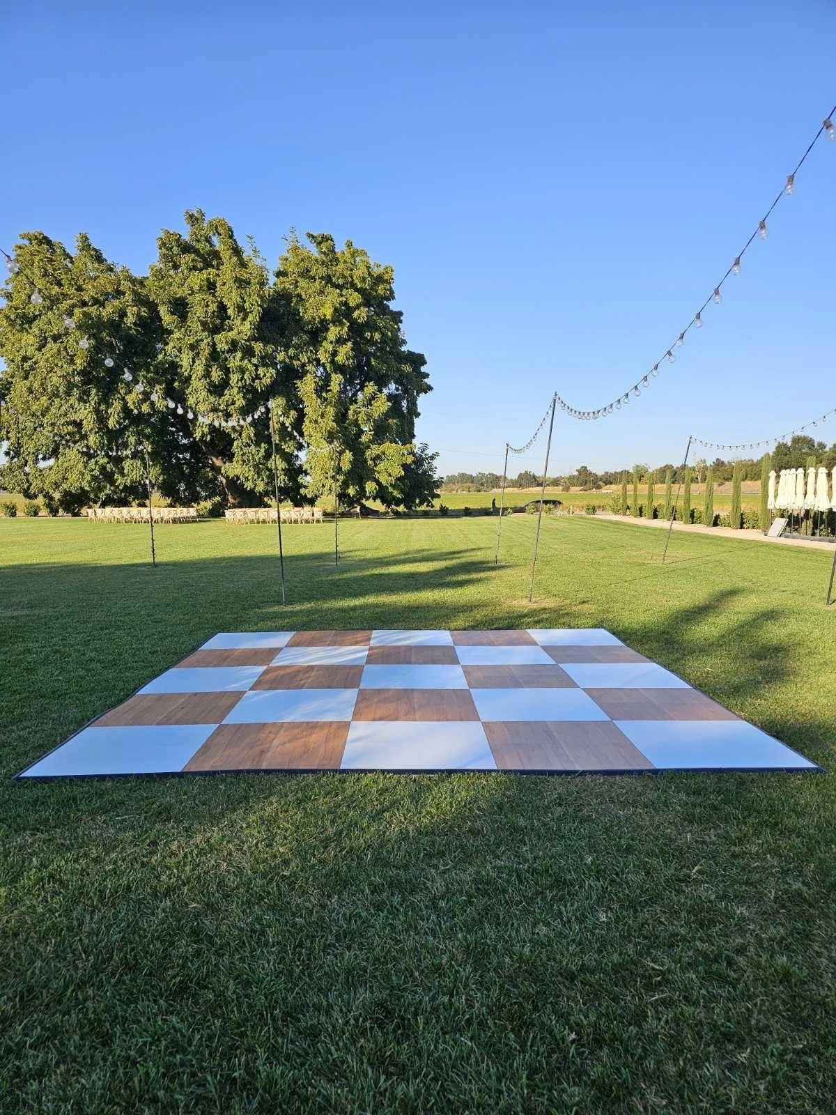 Outdoor dance floor on a grassy lawn with hanging string lights and trees in the background, under a clear blue sky.