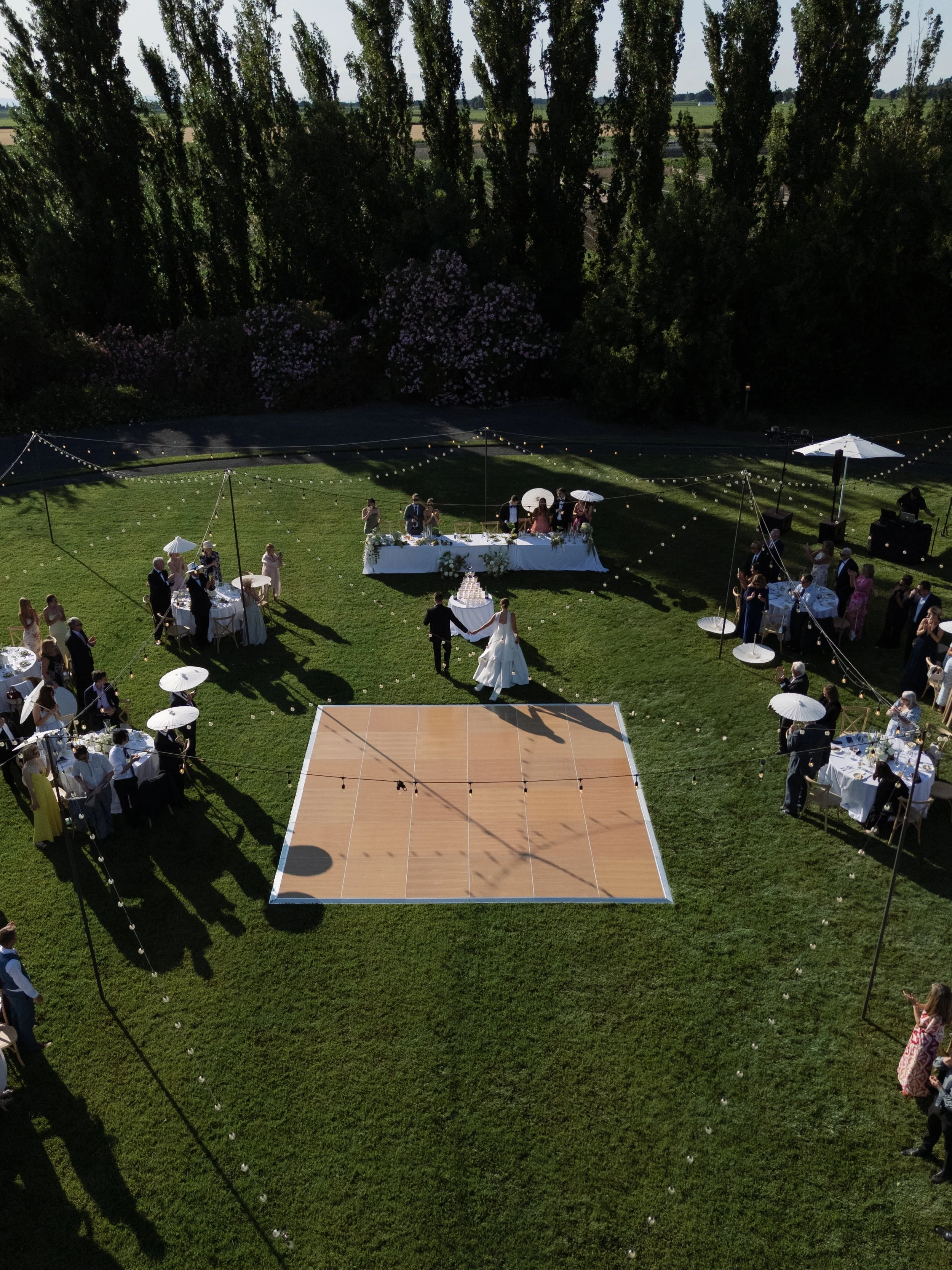 Wedding reception outdoor with tables, guests, string lights, and a dance floor on a grassy area, surrounded by trees.