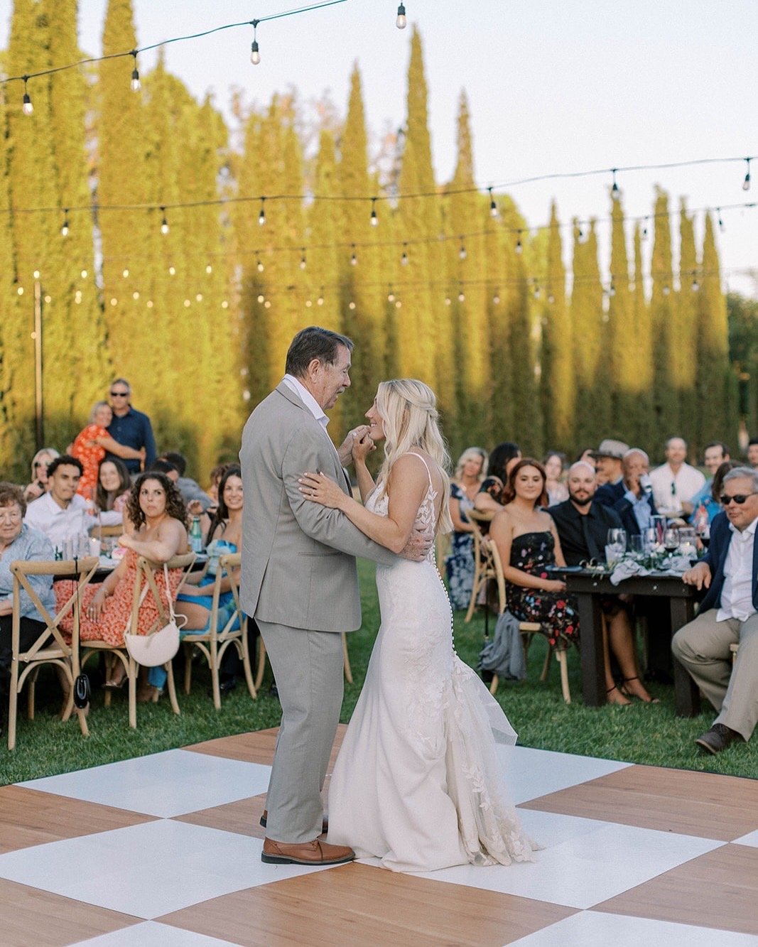 A bride and an older man dancing on a patterned dance floor at an outdoor wedding reception with guests seated at tables and string lights overhead.