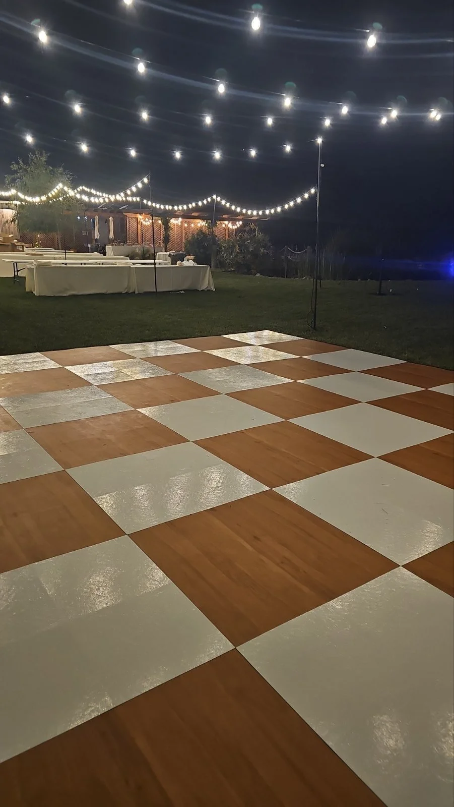 Outdoor event space at night with string lights, tables with white tablecloths, and a multi-colored checkered dance floor.