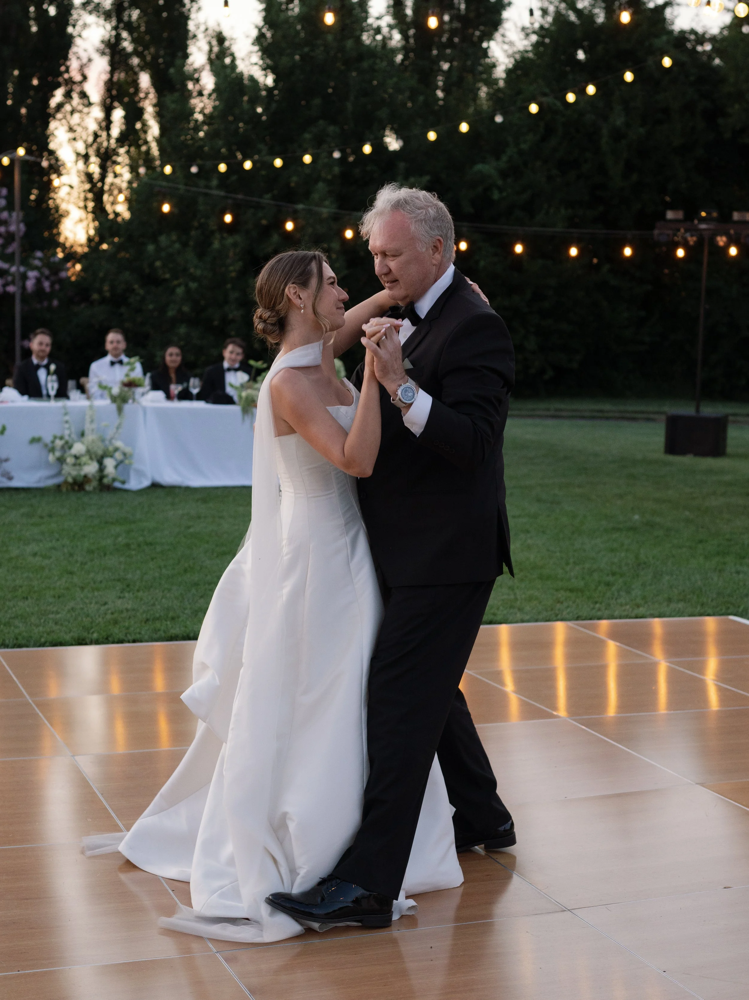 A bride and an older man, possibly her father, dancing at an outdoor wedding reception during sunset with string lights overhead. The bride wears a white wedding gown and the man wears a black tuxedo, both smiling.