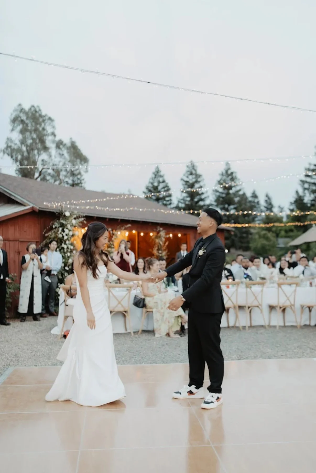 Bride and groom dancing at their outdoor wedding reception with guests watching and string lights overhead.