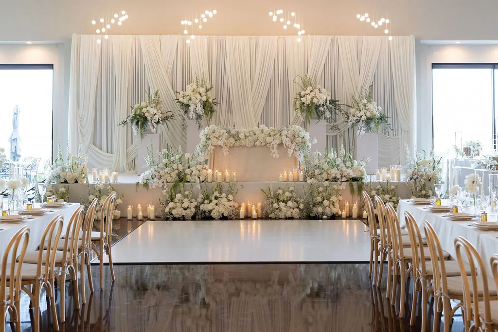 Elegant wedding reception setup with white drapery, floral arrangements, candles, and a dance floor in the center.
