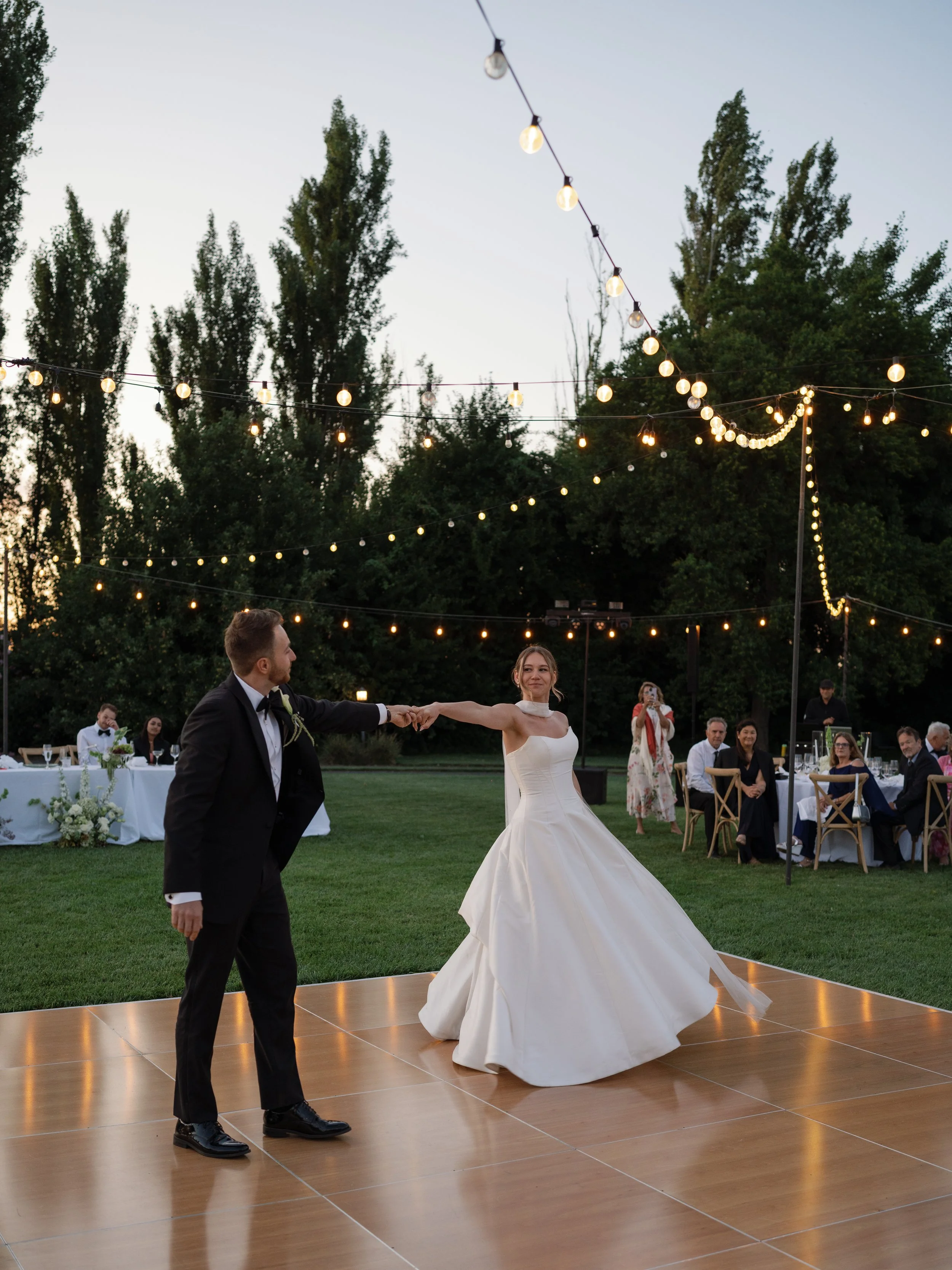 A bride and groom dancing outdoors at their wedding reception, with guests seated at tables in the background and string lights overhead at dusk.