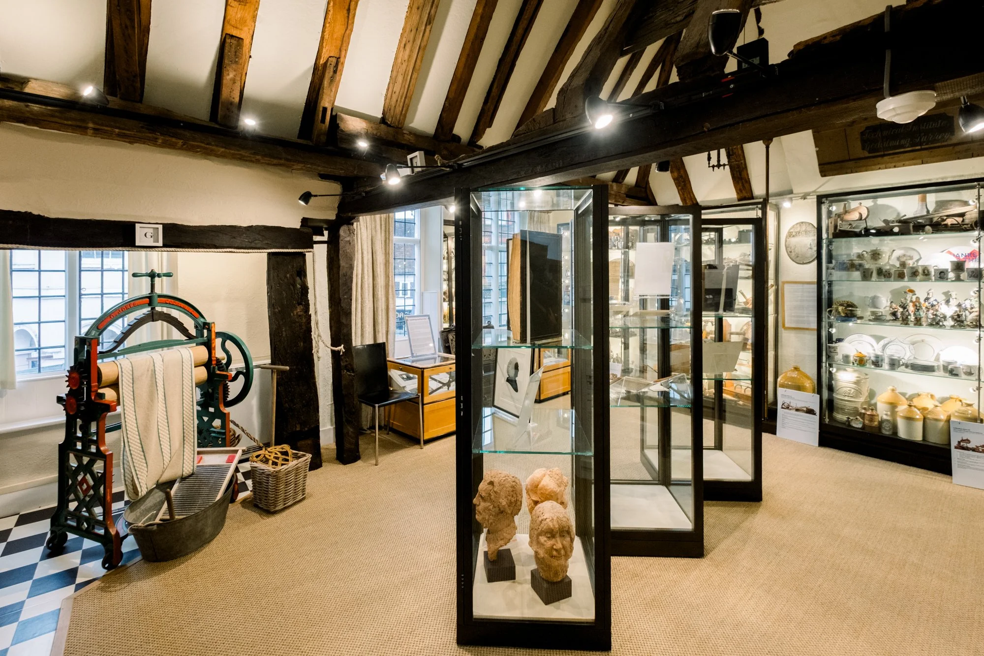 Interior of a museum or exhibit space featuring display cases with artifacts, sculptures, and small collectibles, wooden beams on the ceiling, a window with curtains, and vintage decor items.