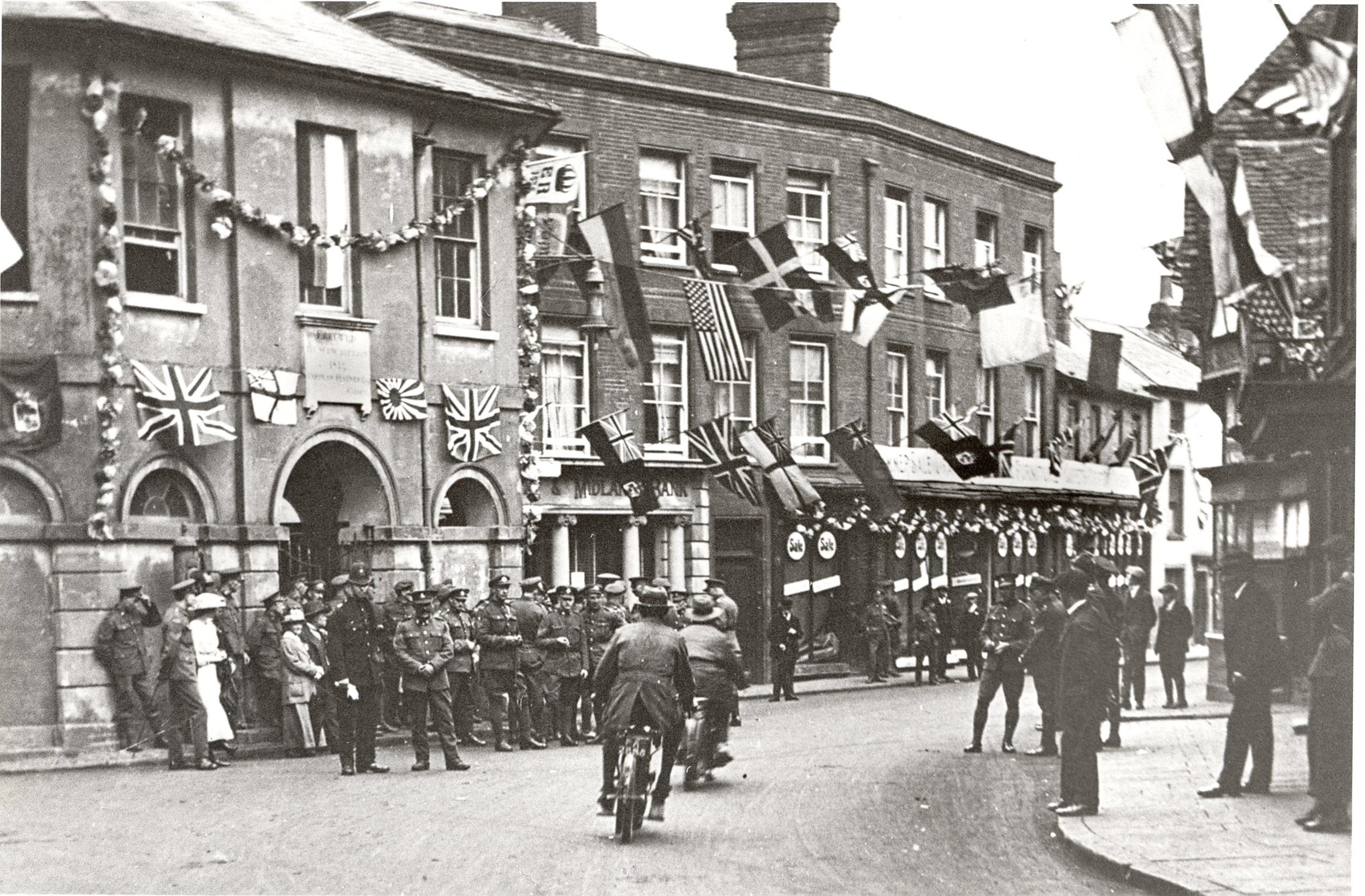 Black and white photo of a street scene with a line of soldiers in uniform standing on the sidewalk and bicyclists riding past. Decorated buildings with flags and banners, possibly celebrating a patriotic event or national holiday.